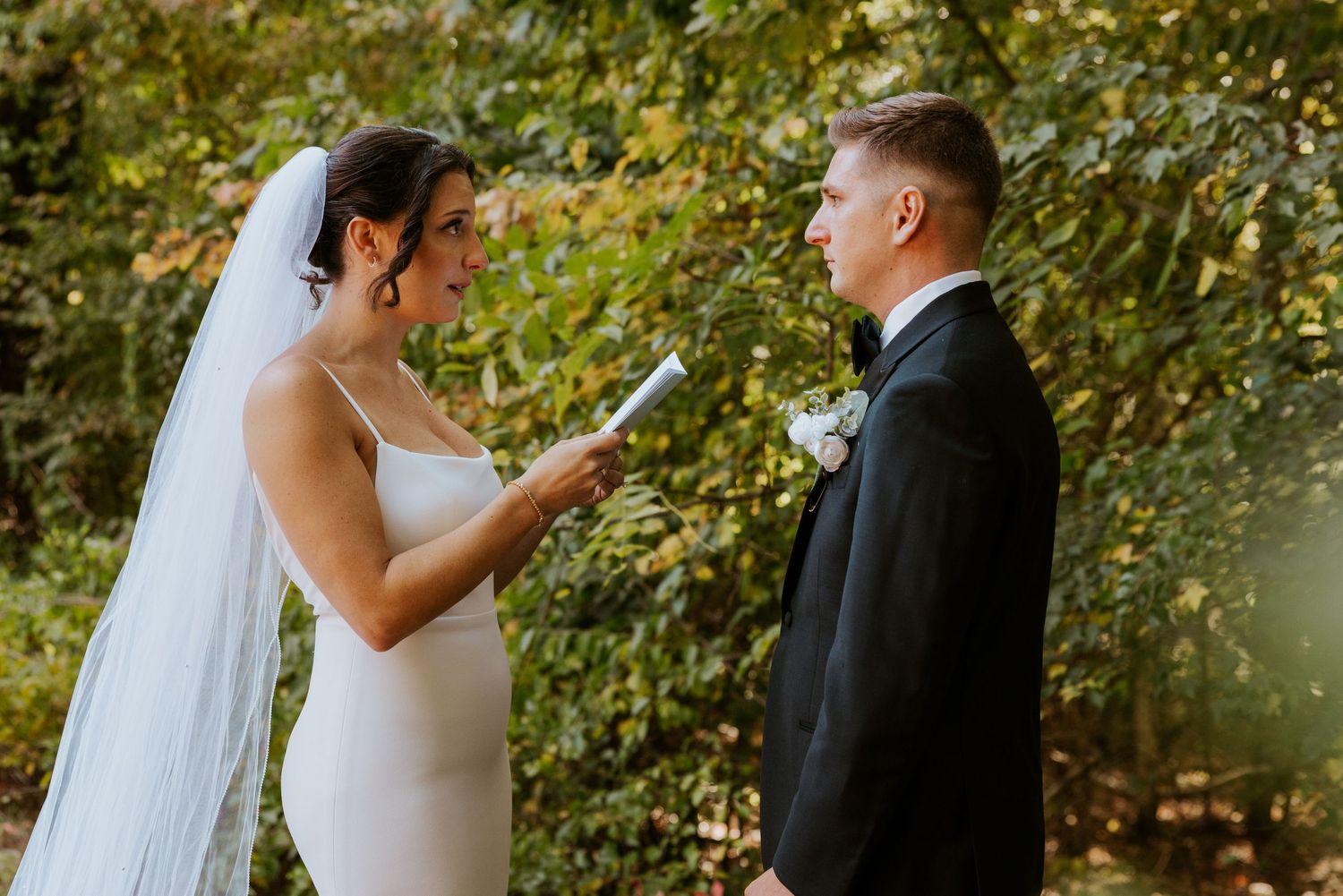 Bride reads wedding vows to groom outdoors in lush green forest setting during intimate ceremony.