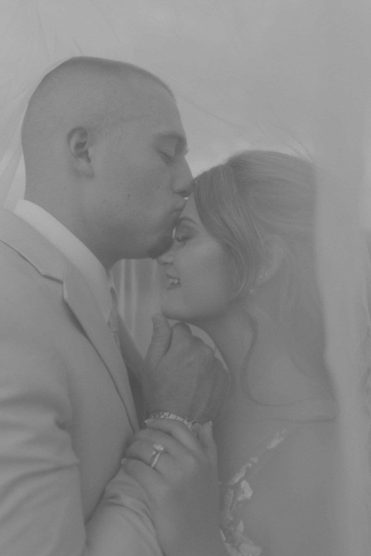 Black and white photo of bride and groom sharing a romantic kiss under a wedding veil.