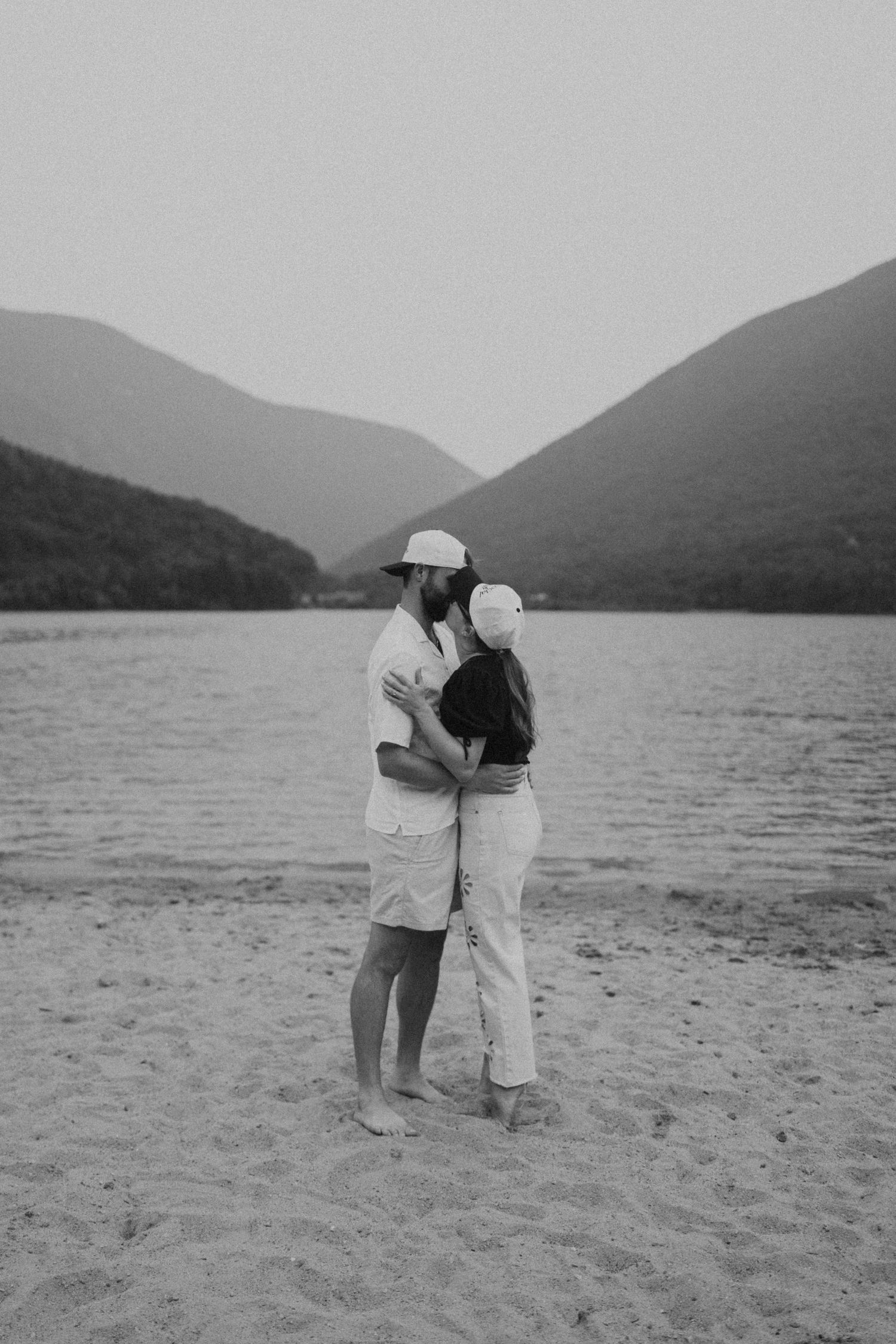 Black and white photo of a couple embracing on a sandy beach with mountain lake views.
