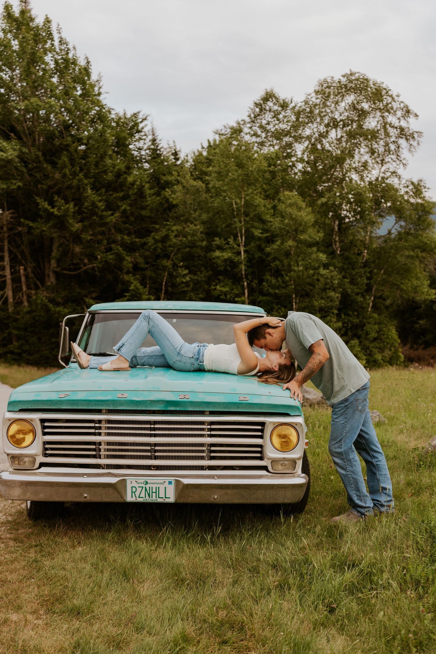 Couple sharing a romantic kiss on the hood of a vintage teal Ford truck in a green field.