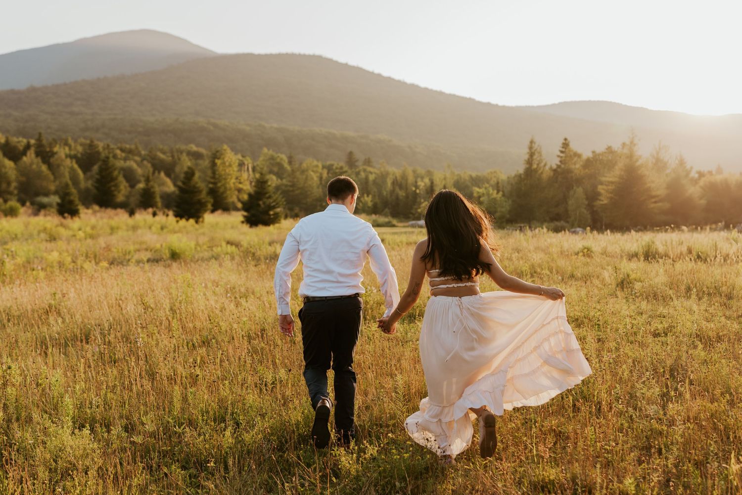 Couple runs hand in hand through golden field toward stunning mountain landscape.