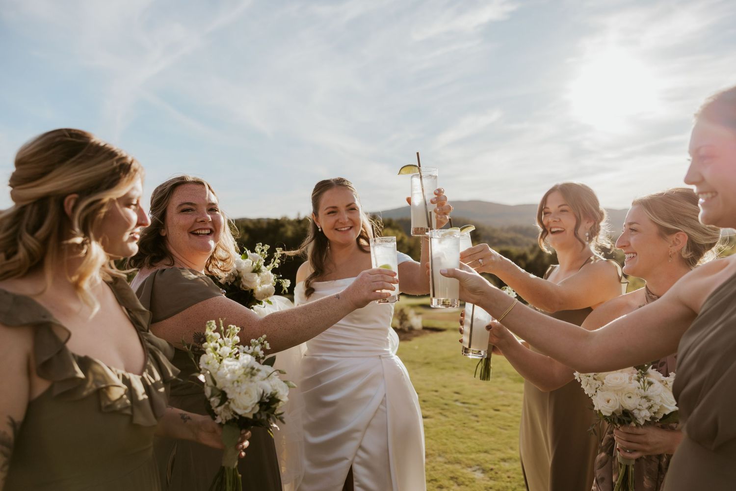 Bride celebrates with bridesmaids in olive dresses toasting with drinks at outdoor wedding reception.