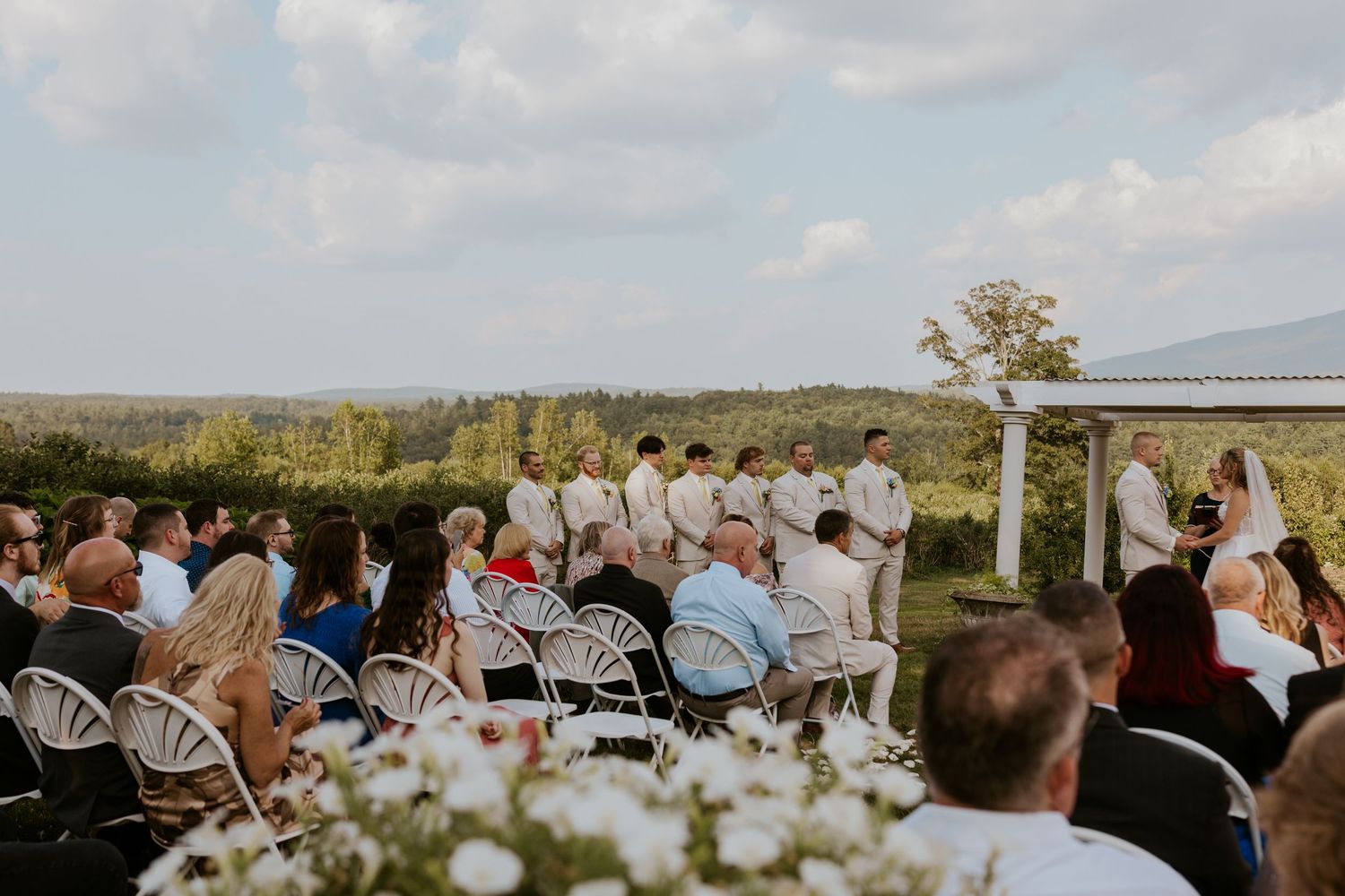Outdoor wedding ceremony with guests seated in white chairs overlooking scenic vineyard landscape under blue sky.