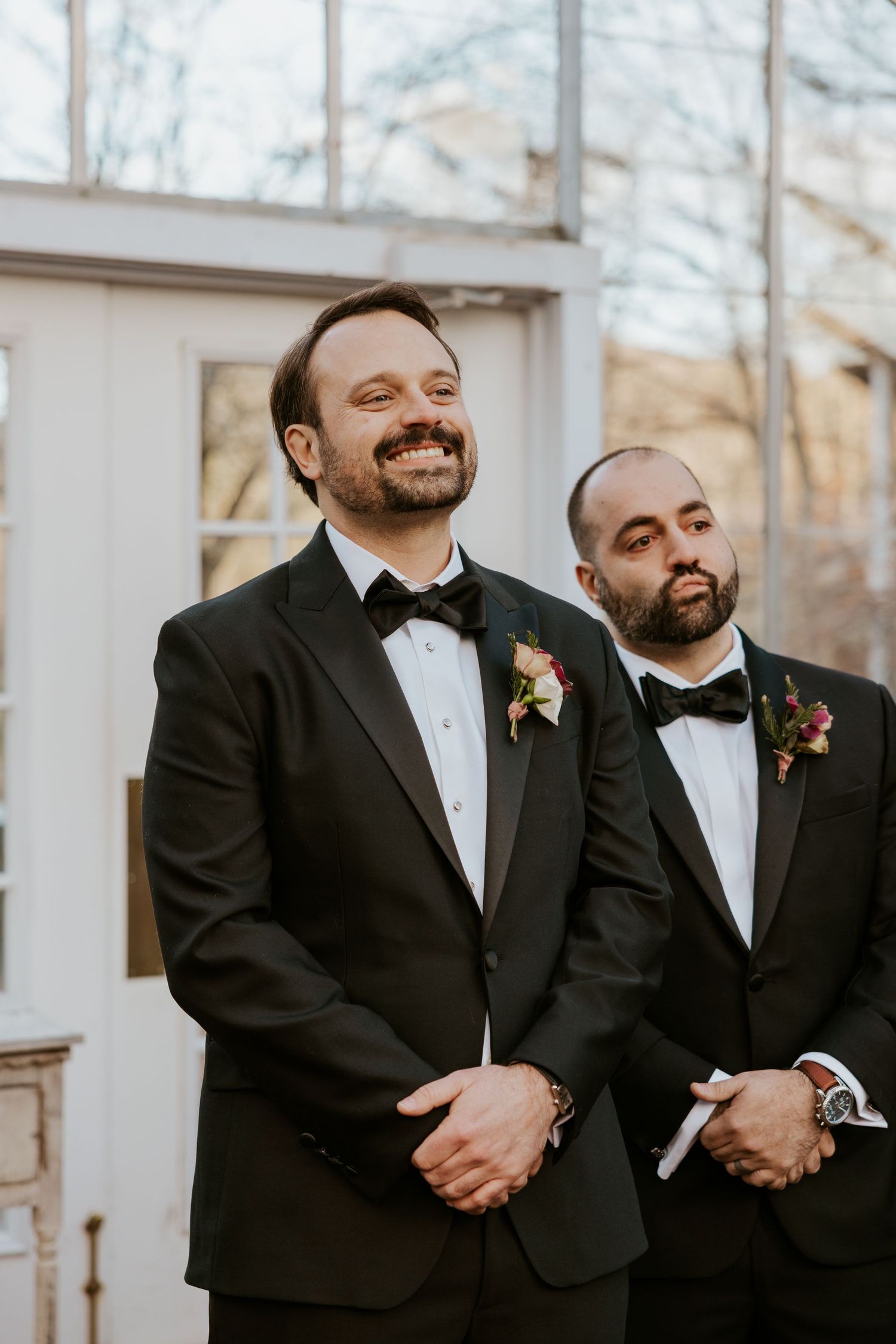 Smiling groom and best man in black tuxedos with floral boutonnieres await the bride at the altar.