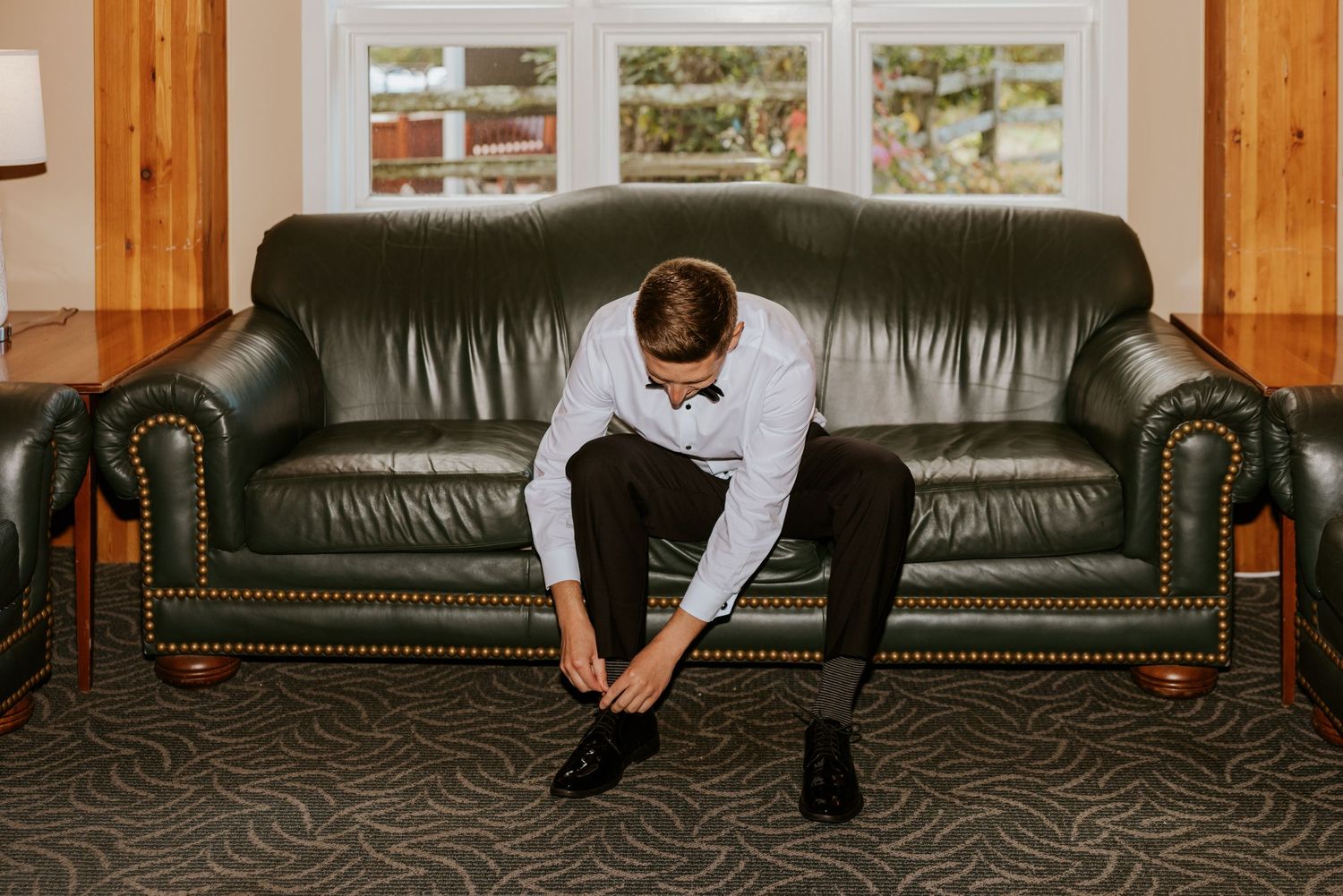 Groom sits on green leather sofa tying his dress shoes before the wedding ceremony.