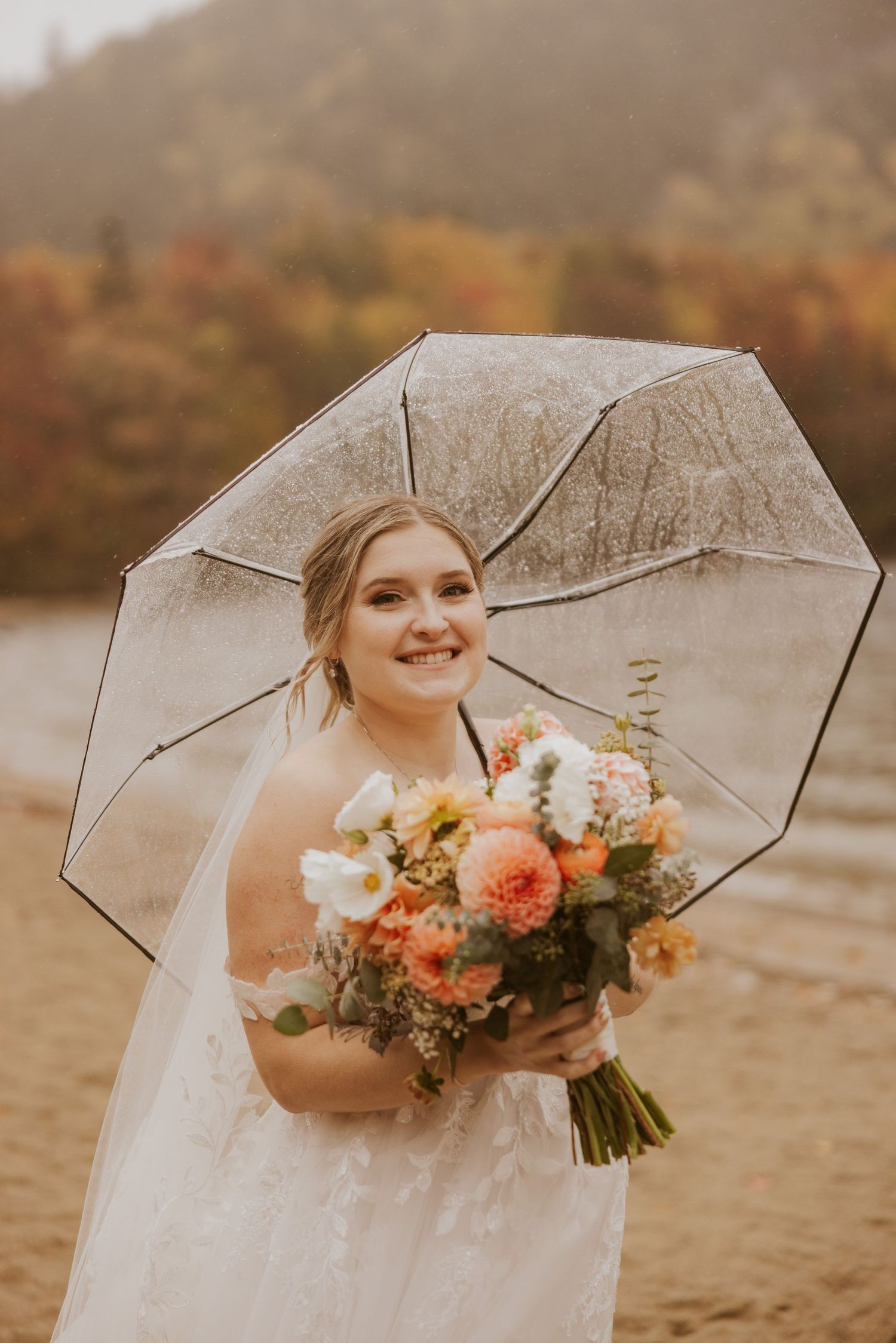 Radiant bride holds a lush peach and white bouquet under a clear umbrella on a rainy fall day.