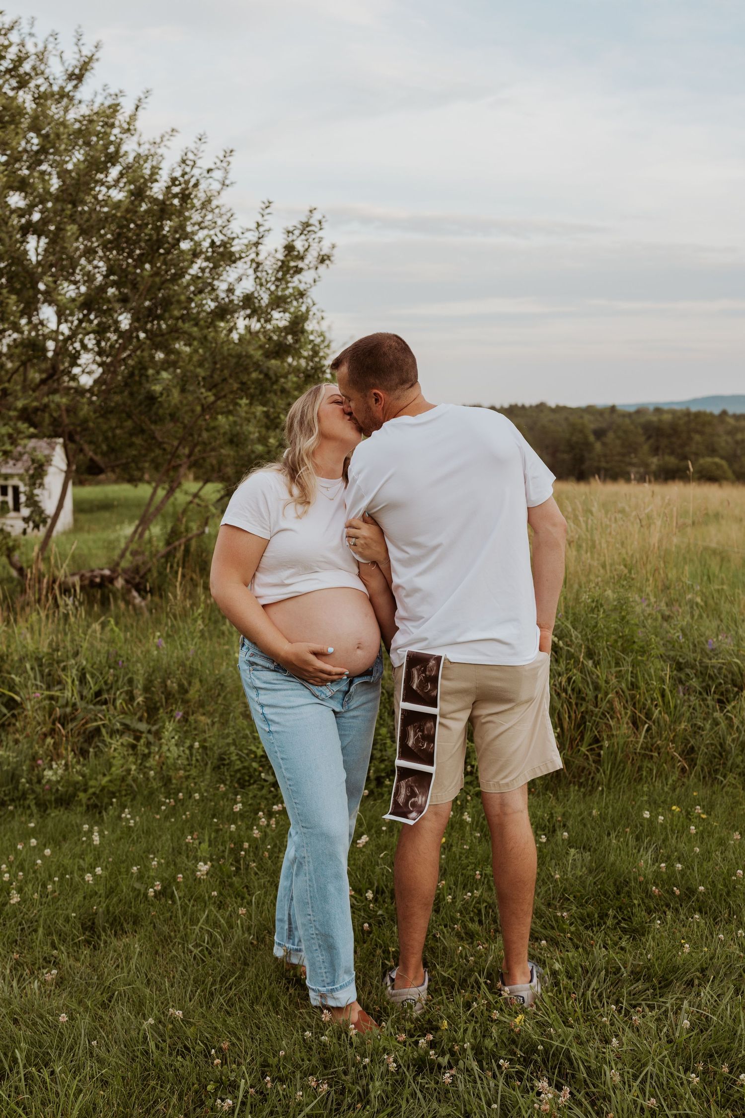 Pregnant woman and partner kiss in sunny meadow while man holds ultrasound photos at his side.