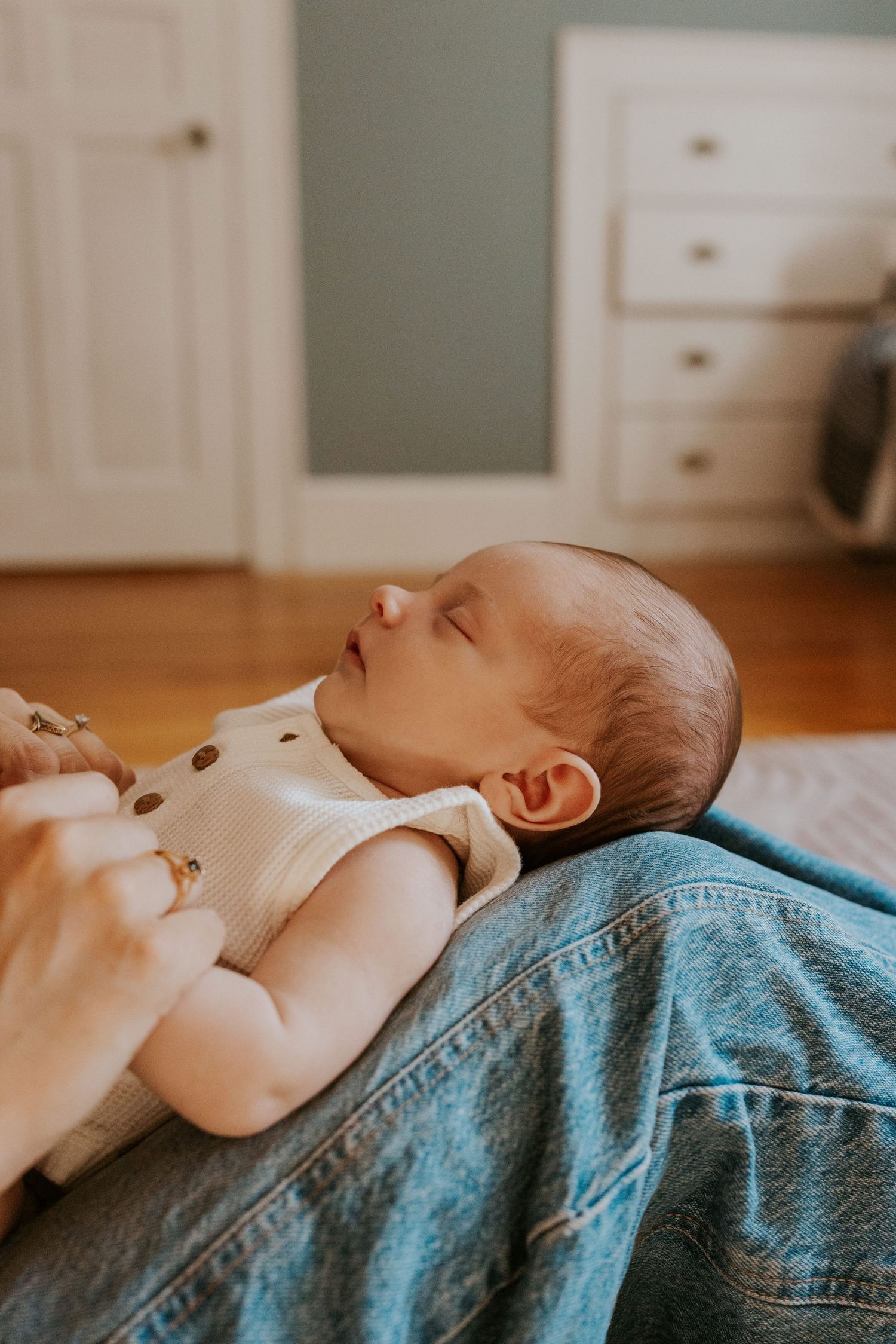Sleeping newborn baby in cream onesie resting on parent's denim-clad legs on hardwood floor.