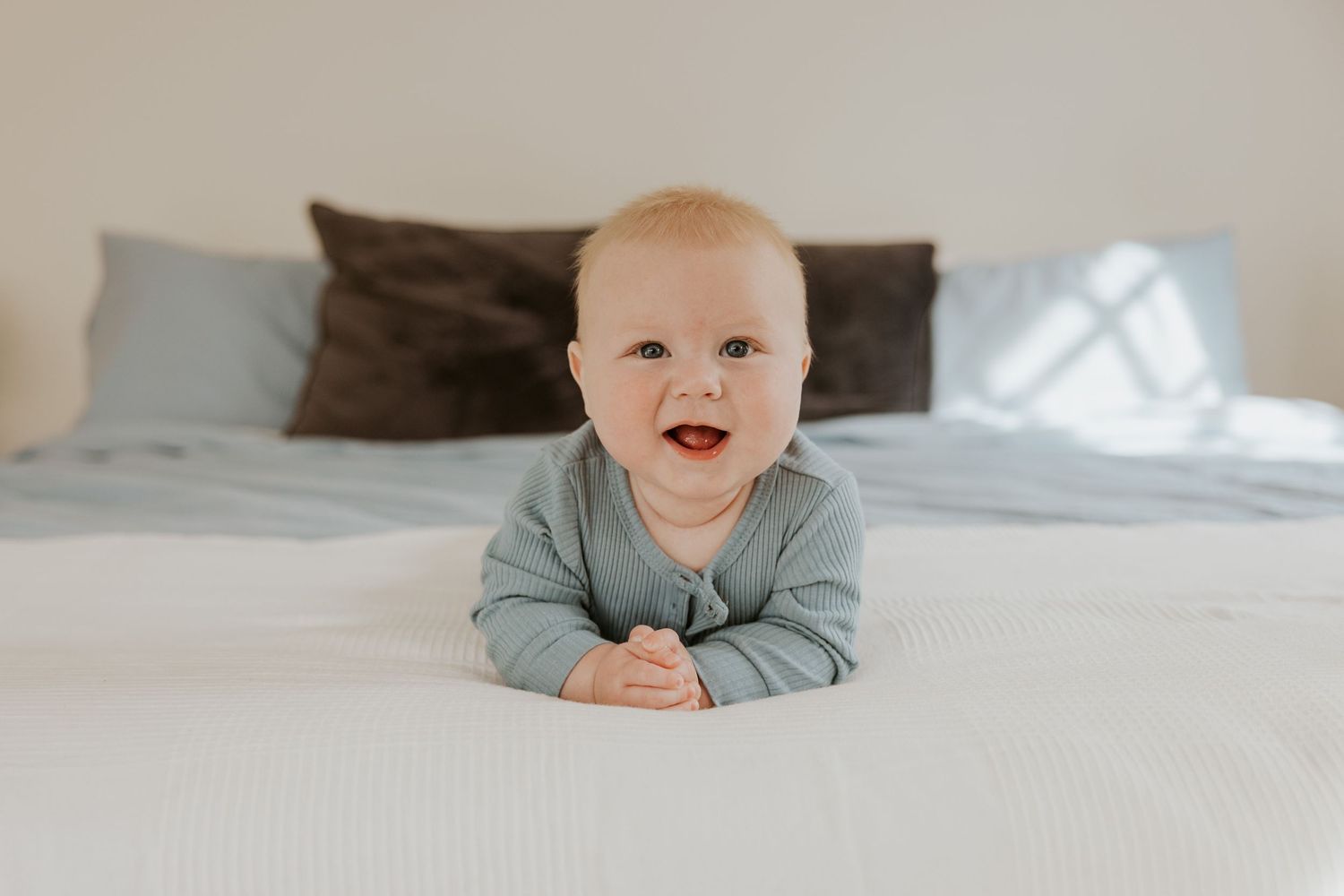 Adorable smiling baby in sage green outfit doing tummy time on a white bed.