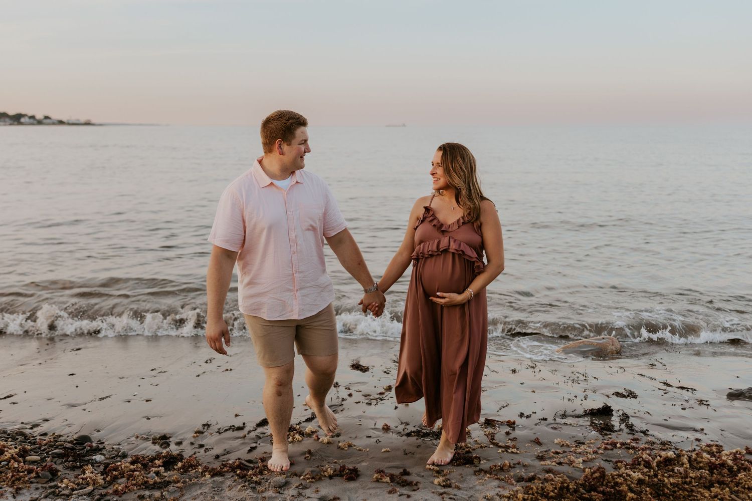Pregnant couple holding hands walking along rocky shoreline at sunset in romantic maternity photo.
