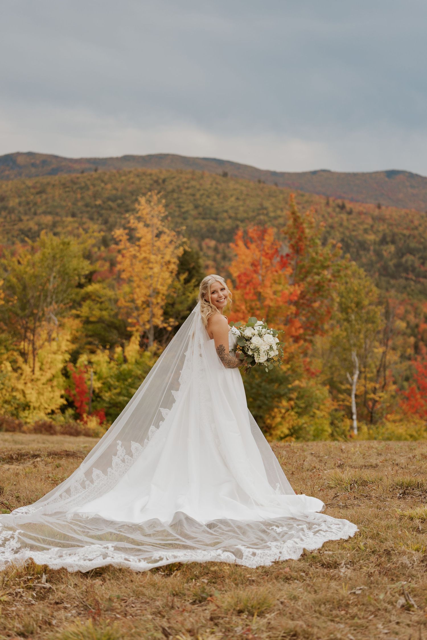 Bride in white gown with long lace veil holds bouquet in autumn mountain landscape with colorful foliage.