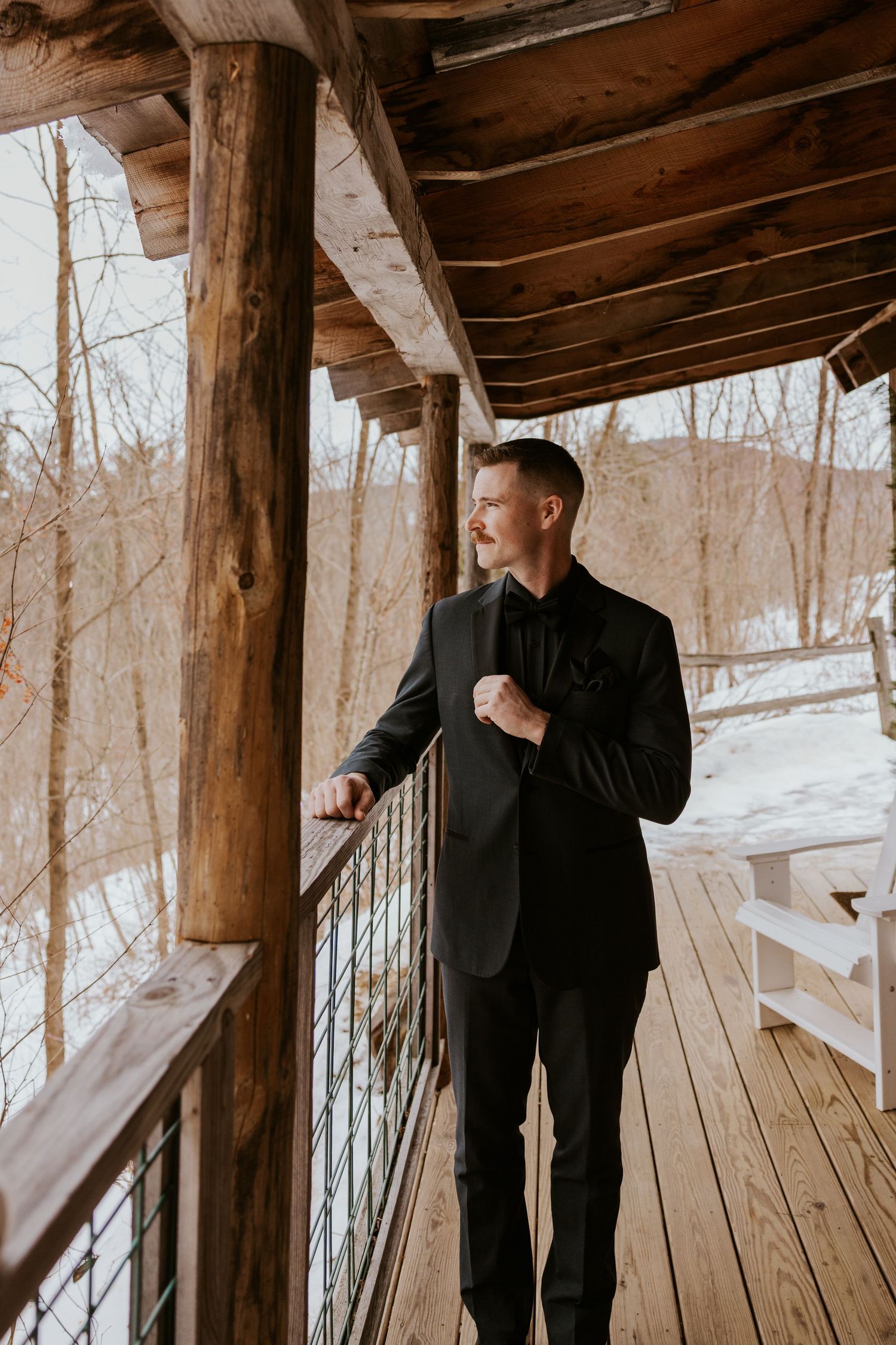 Groom in black suit standing on rustic wooden porch overlooking snowy winter landscape.