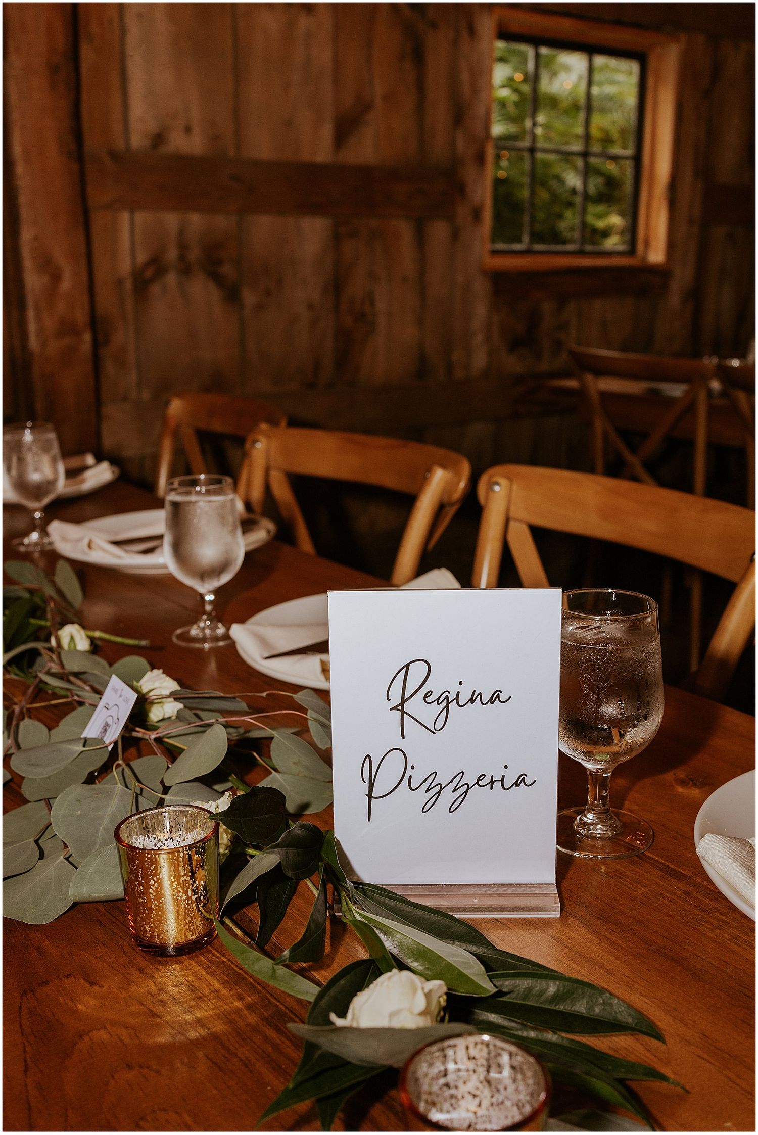 Rustic wedding reception place cards with greenery and eucalyptus on wooden tables with glassware and candlelight.