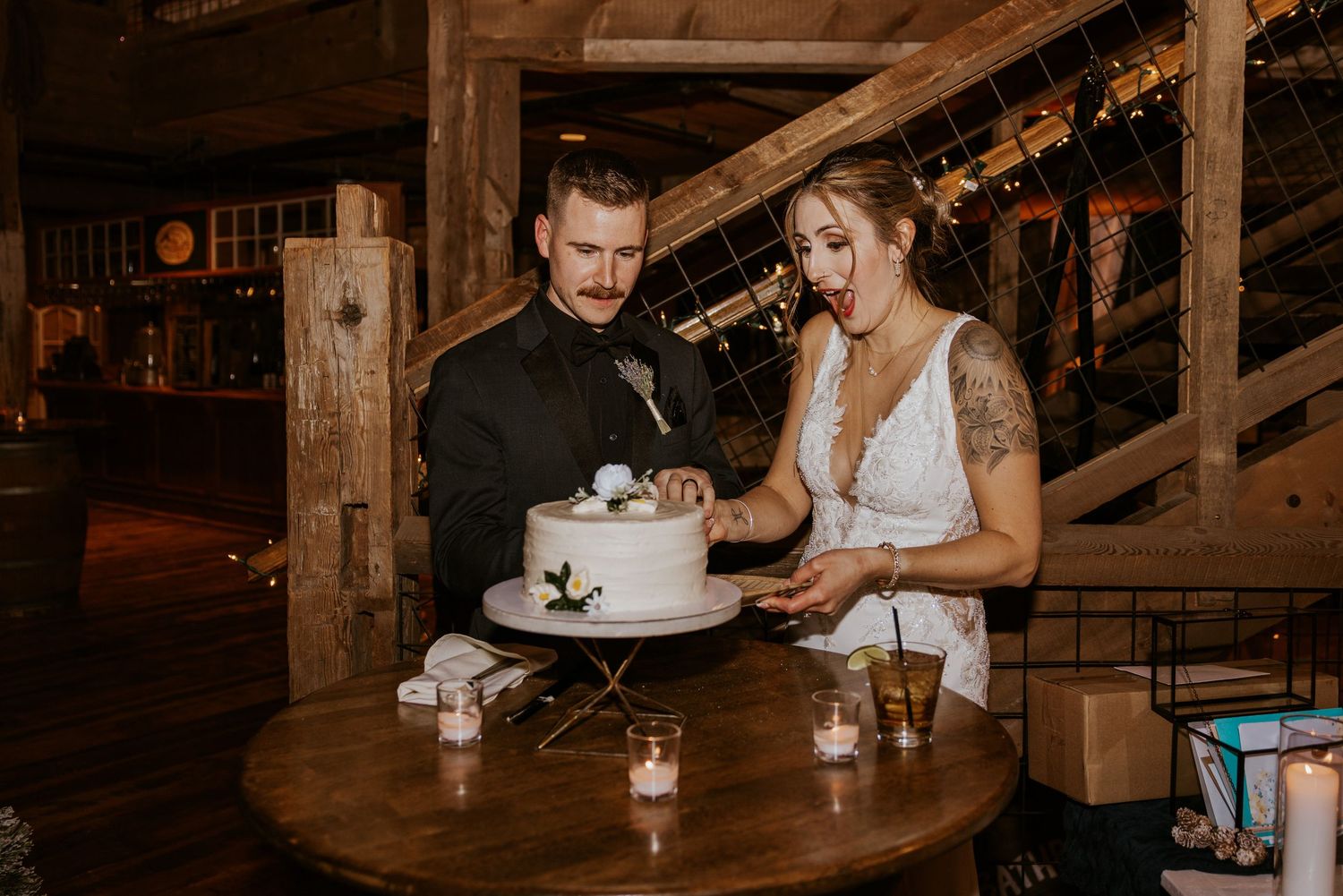 Bride and groom cutting their white wedding cake at a rustic barn venue with warm candlelight ambiance.