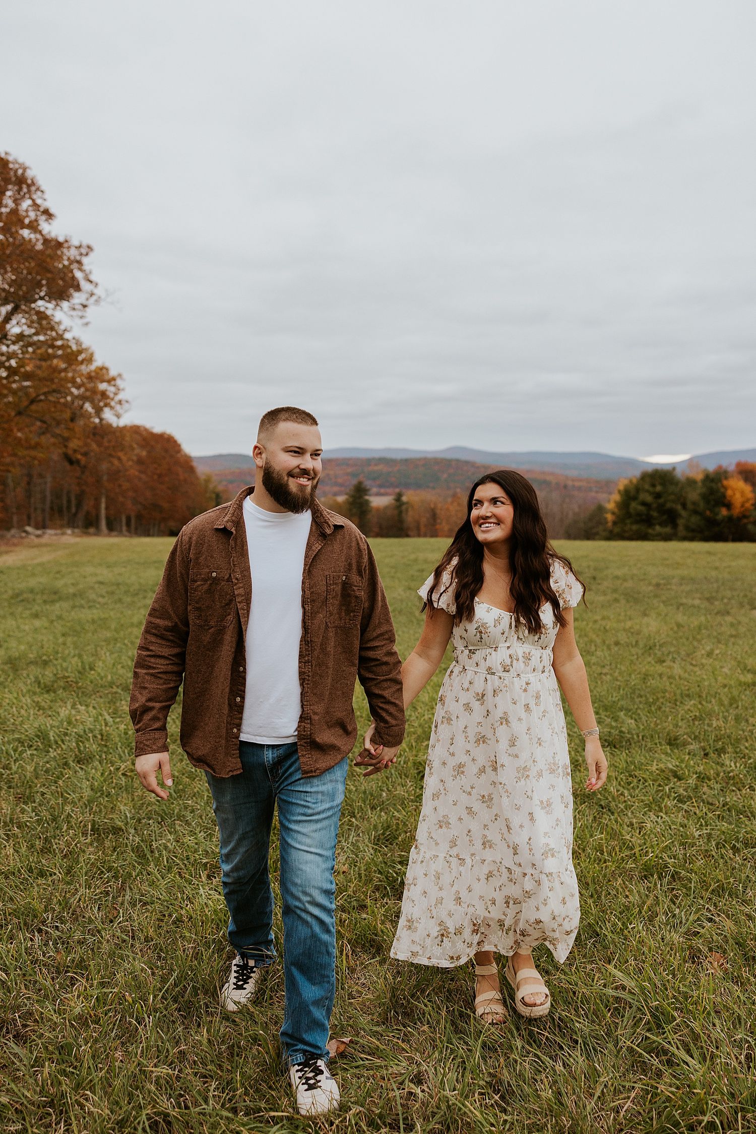 Couple holding hands walking in a green field with autumn trees and mountains in the background.