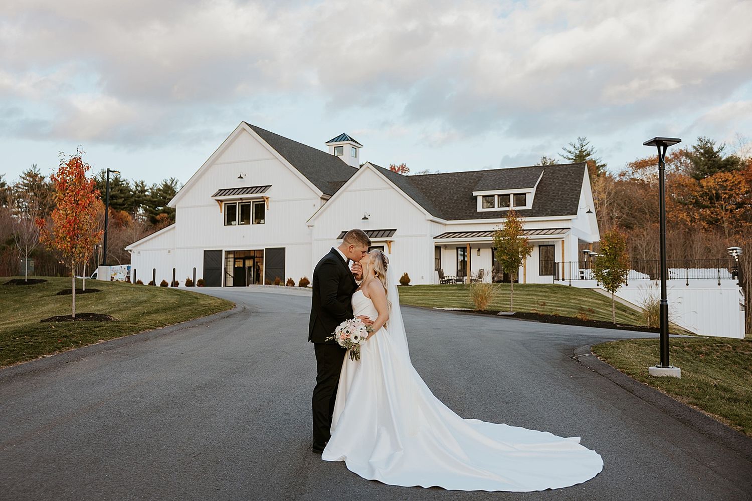 Bride and groom share a kiss on a driveway outside a white barn wedding venue with autumn foliage in the background.