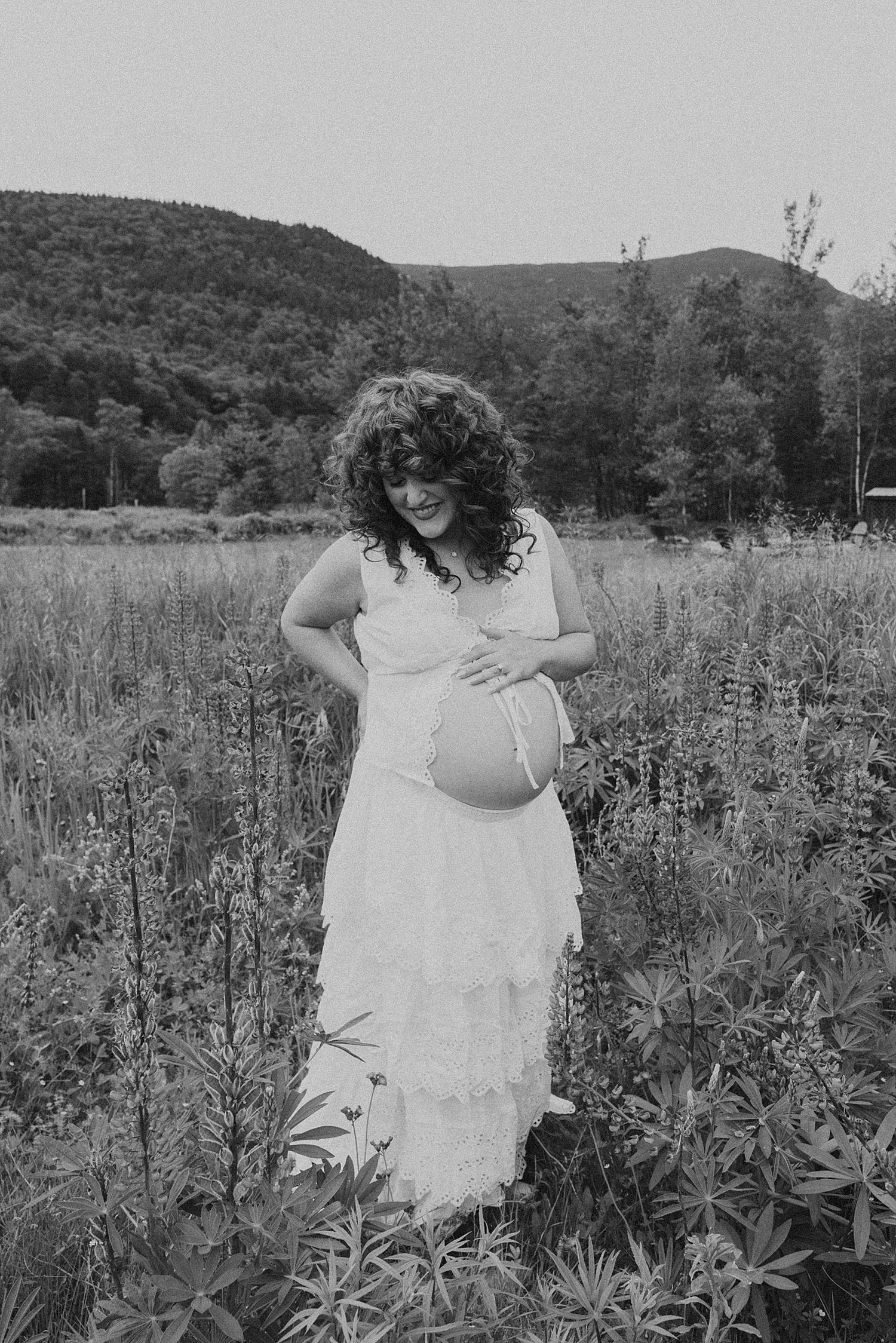 Pregnant woman in white lace dress holding baby bump in a wildflower field, black and white maternity photo.