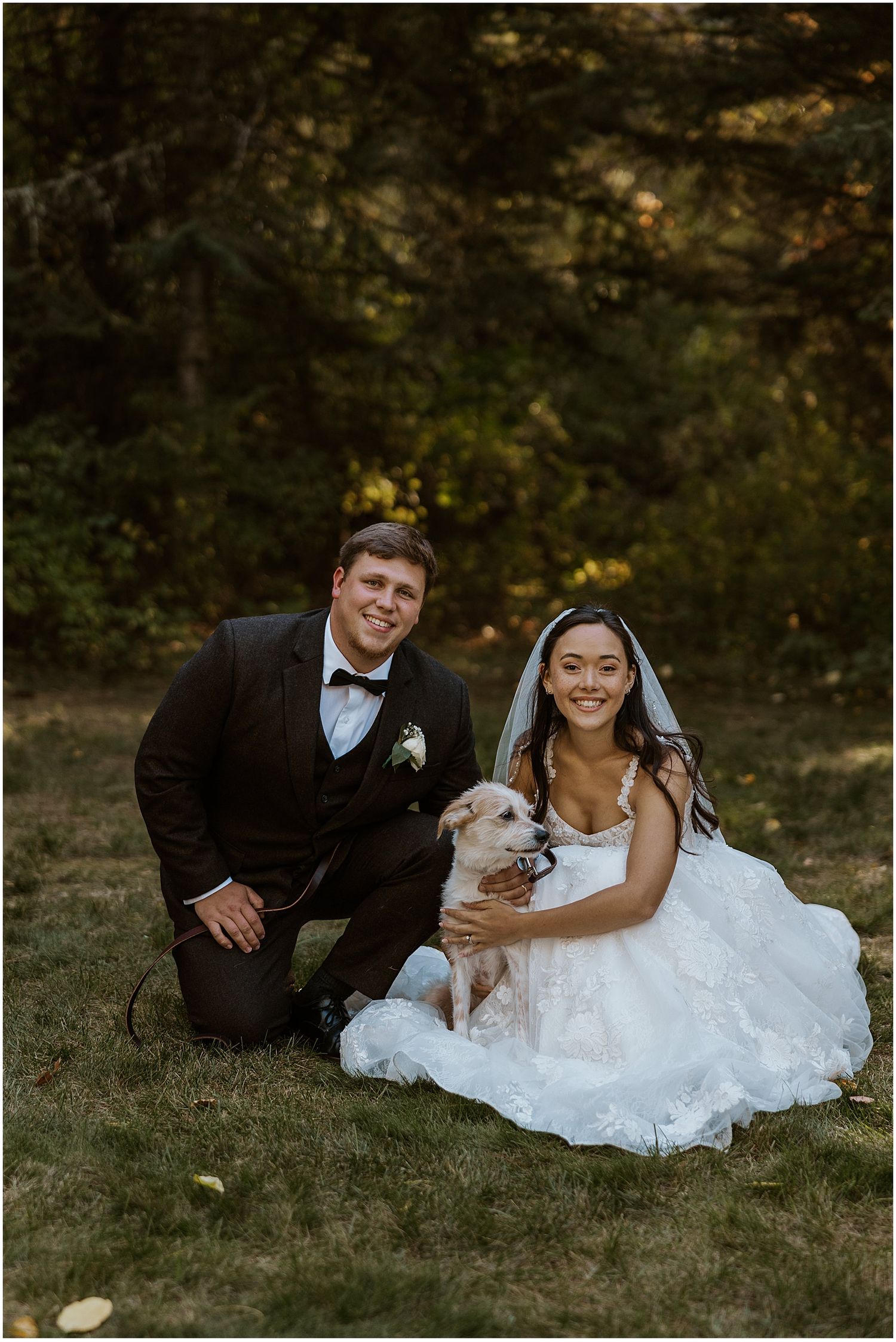 Newlyweds pose with their small dog on grassy lawn during outdoor wedding photoshoot.