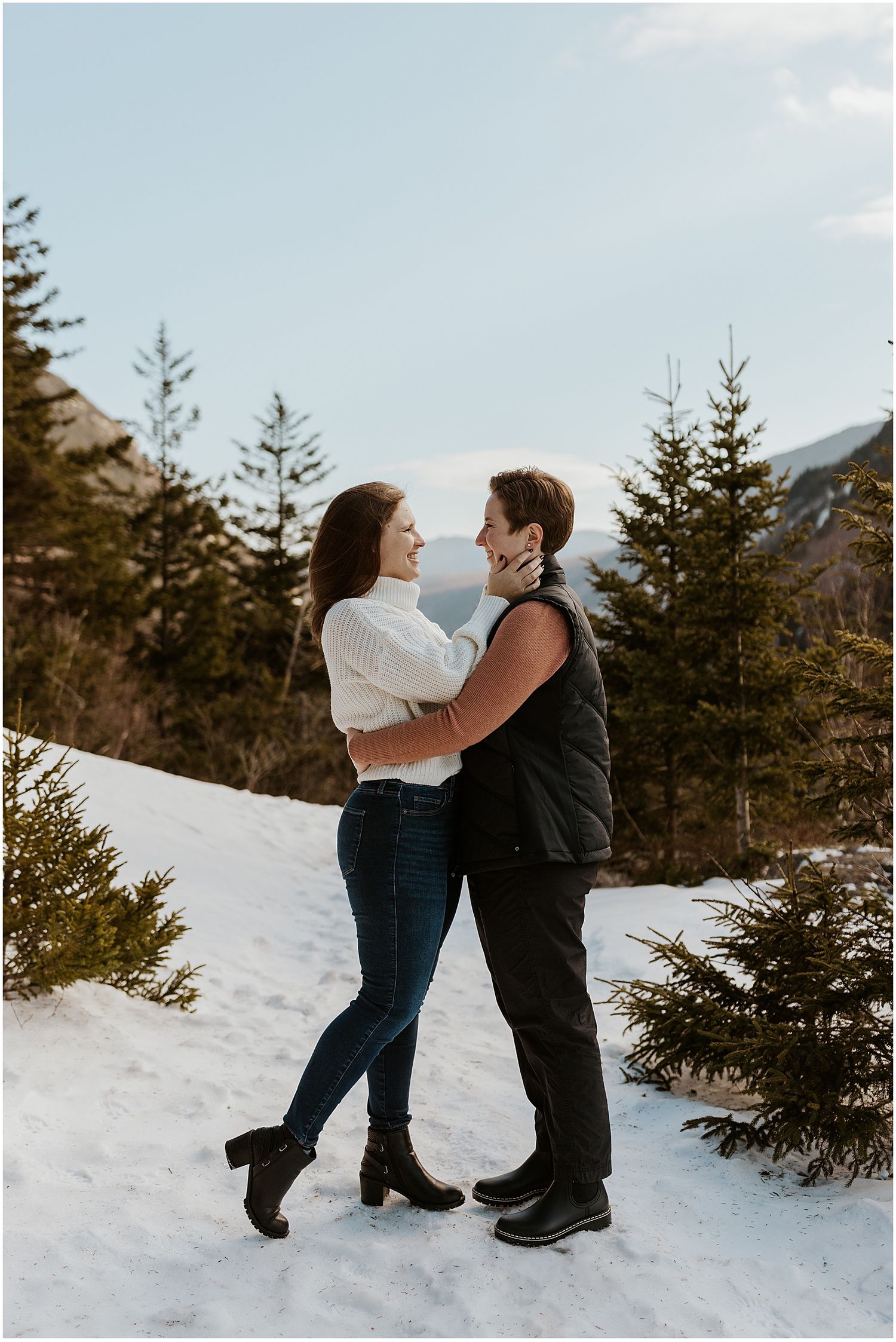 Playful winter engagement photoshoot in the snow with evergreen trees.