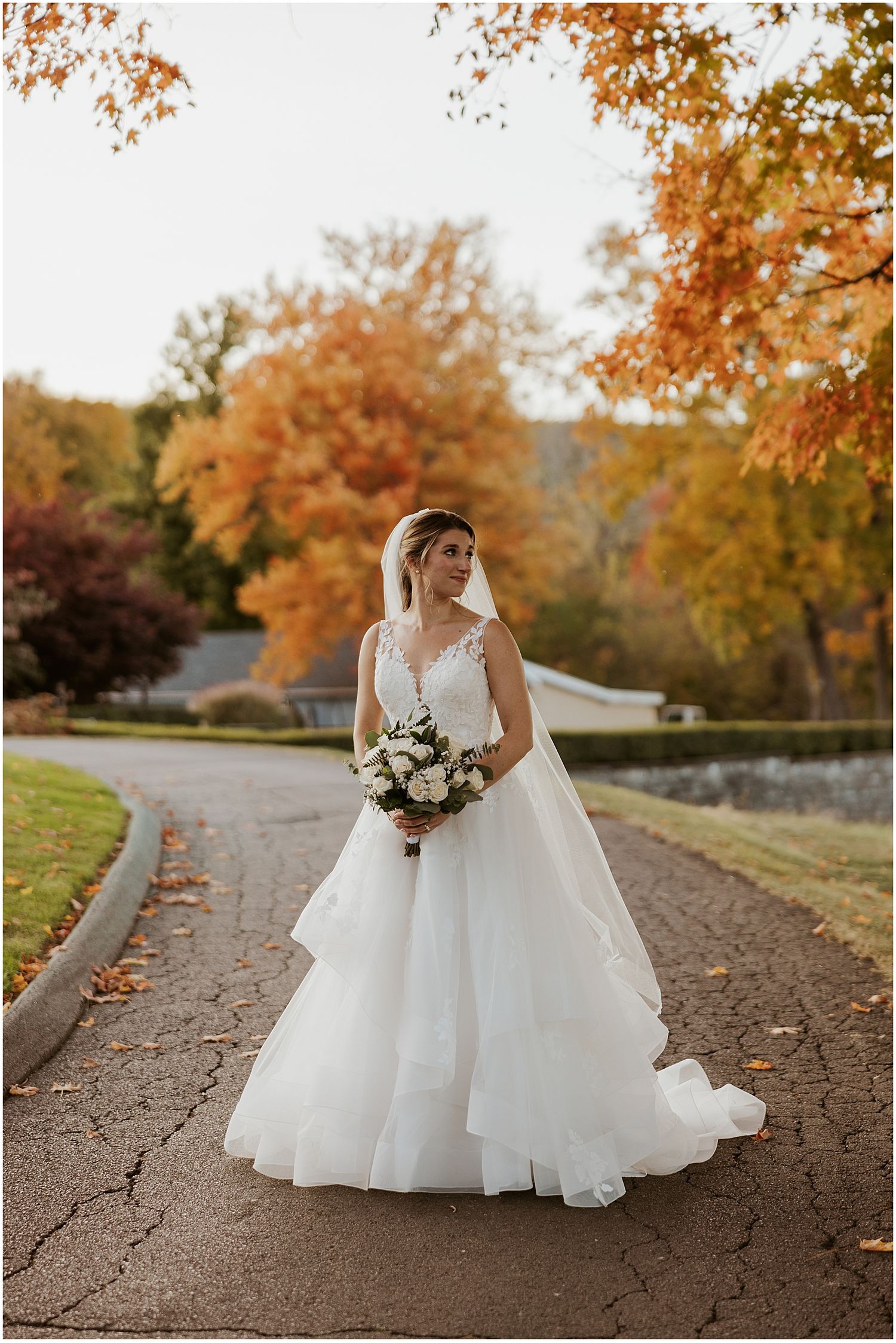 Bride in a flowing white ballgown poses on a path surrounded by vibrant fall foliage.