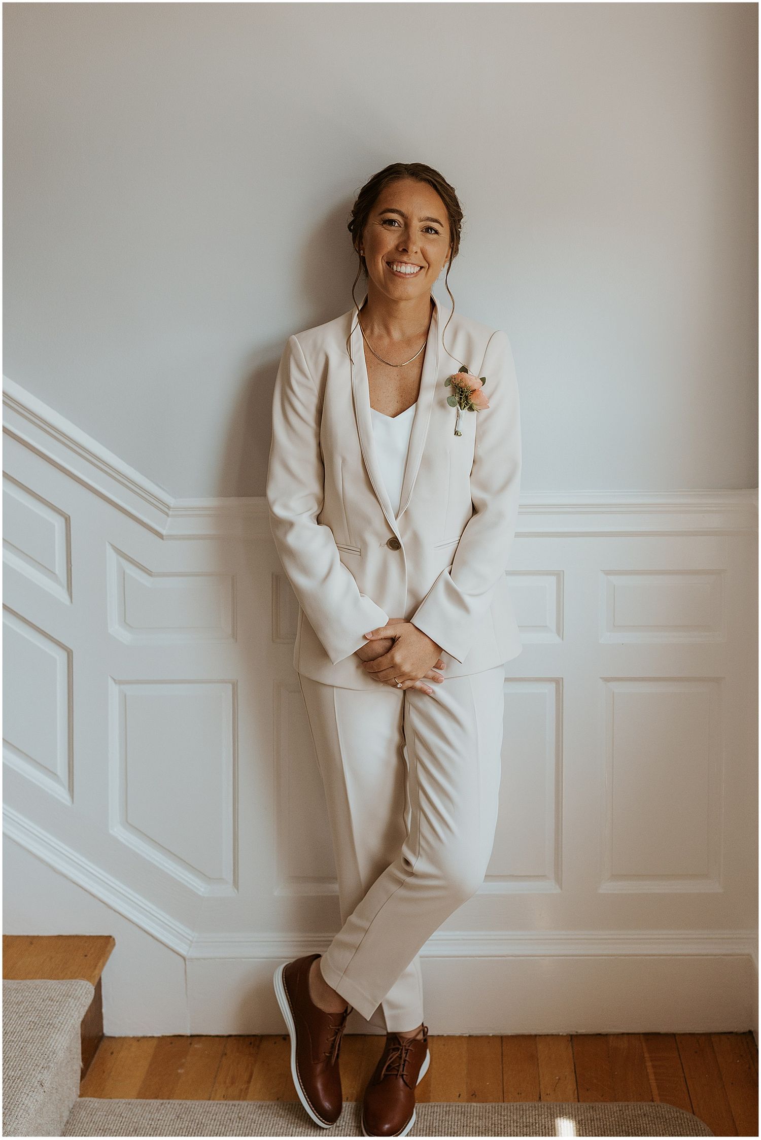 A person wearing an elegant white suit and floral boutonniere poses casually against a paneled wall.