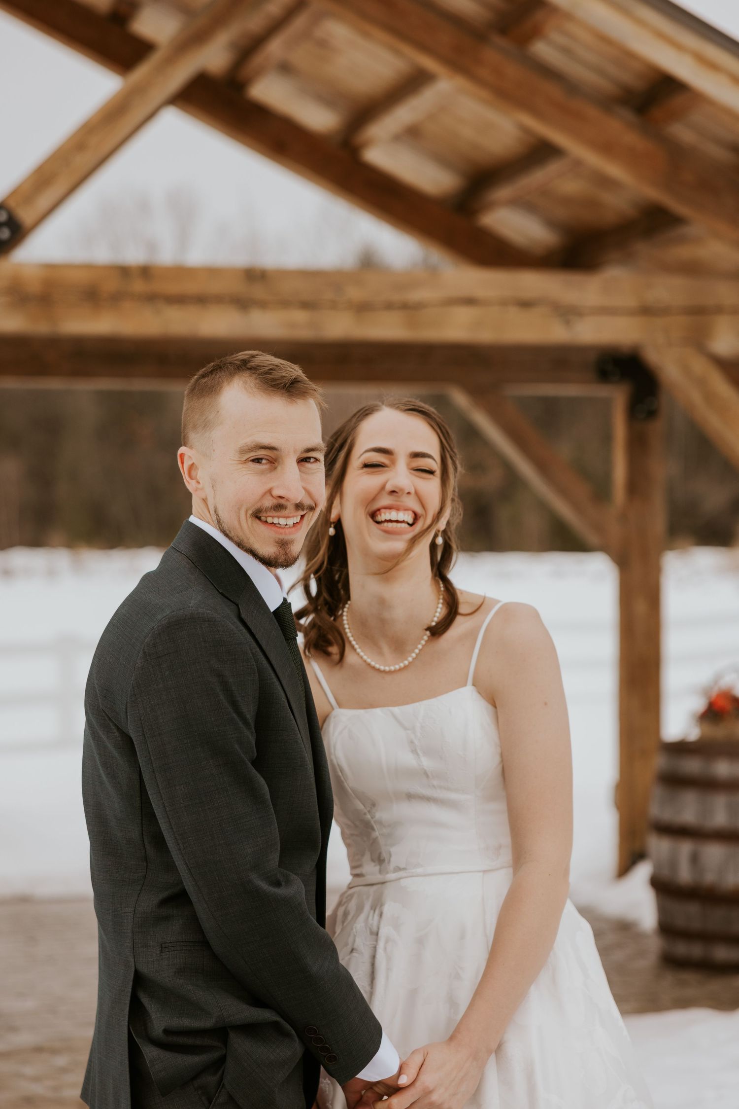 A joyful couple shares a candid moment together under a rustic wooden pavilion during their winter wedding photoshoot.