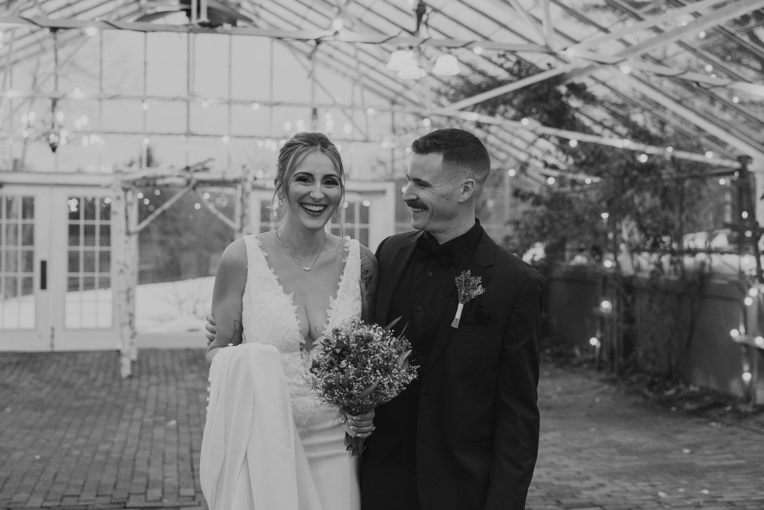 Black and white photo of smiling bride and groom inside a greenhouse wedding venue, bride holding bouquet.
