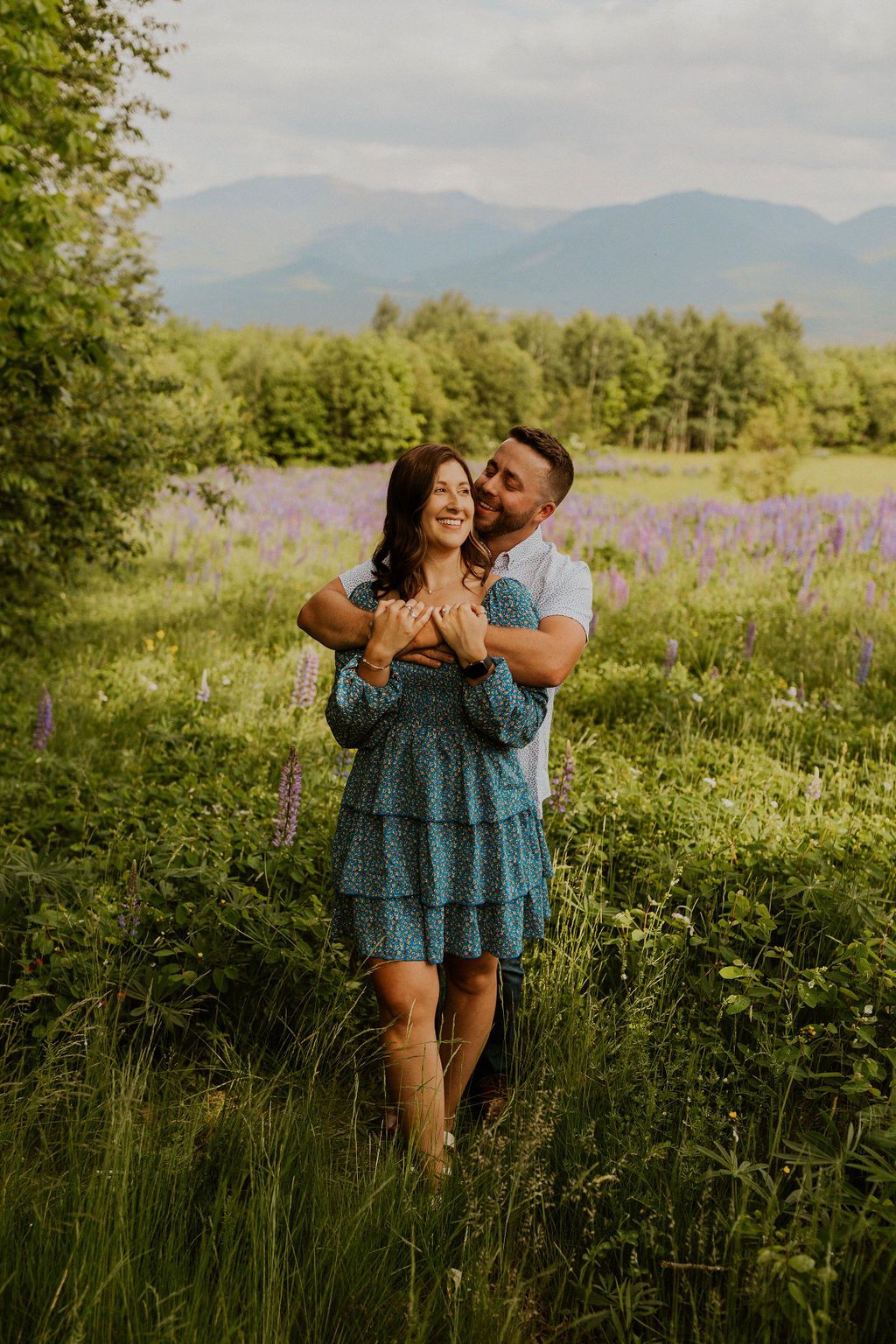 Romantic embrace in a meadow of purple wildflowers with mountains in the background.