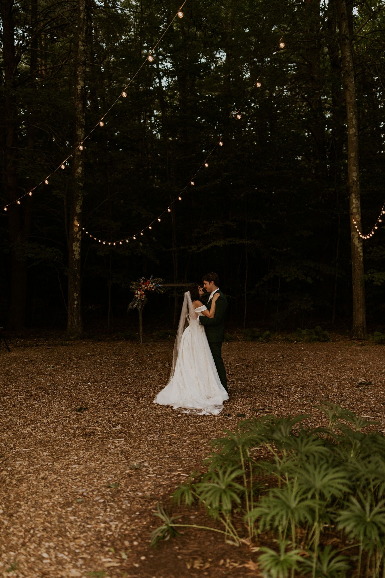 String lights illuminate a romantic wooded outdoor wedding venue at dusk.