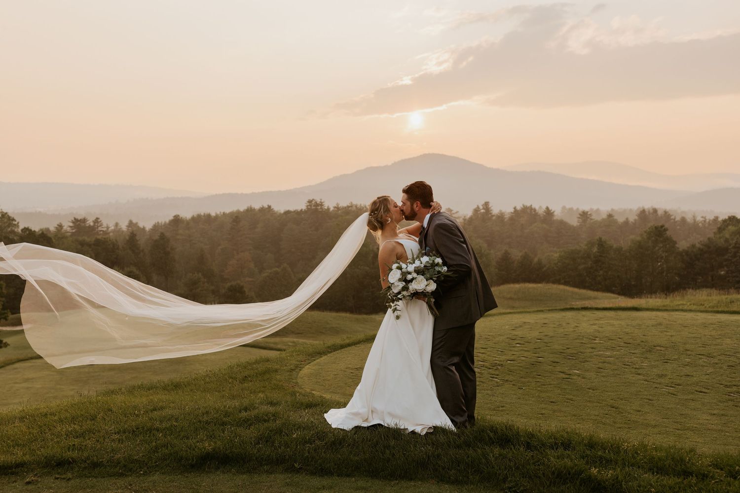 Bride and groom kiss at golden sunset with mountain views and flowing veil in the wind.