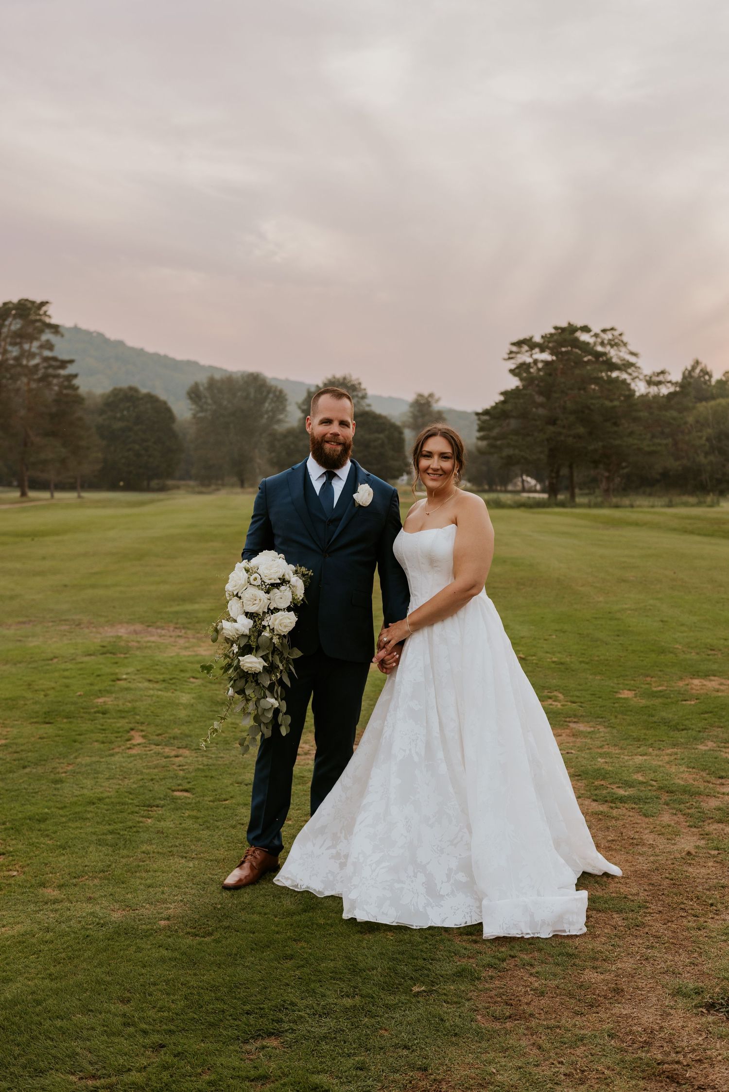 Bride and groom posing together on a golf course at sunset, bride holding a white floral bouquet.