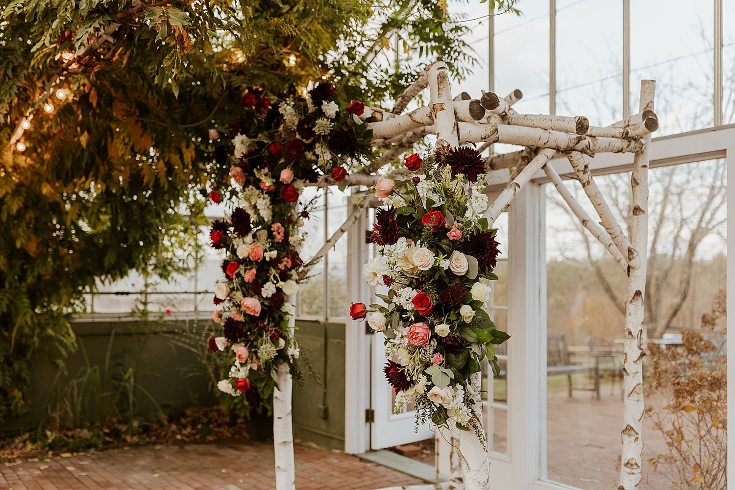 White birch wedding arch adorned with burgundy, blush, and white floral arrangements inside the greenhouse at the Barn on the Pemi NH