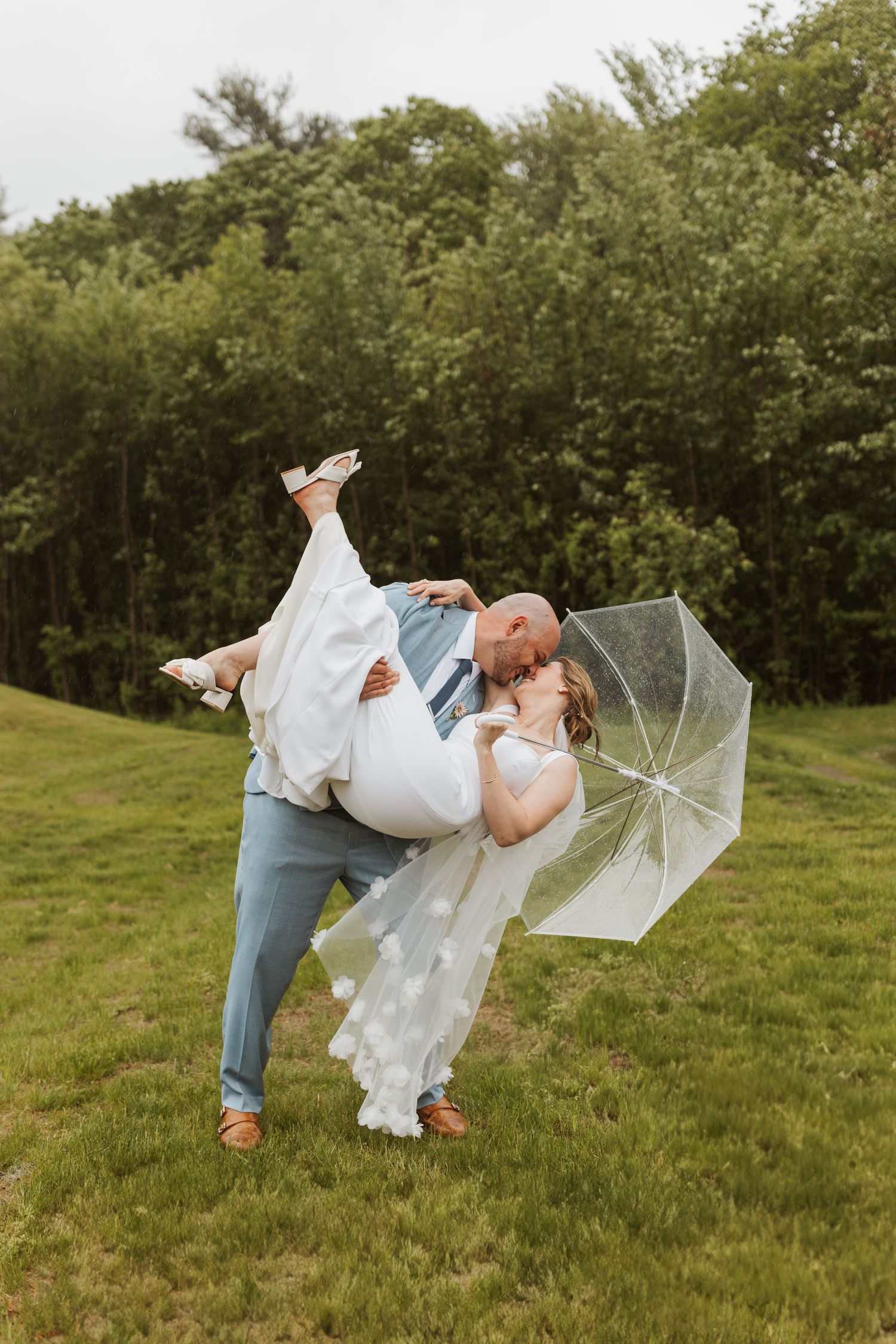 Groom dips bride in a playful kiss on a green lawn while she holds a clear umbrella on a cloudy day.