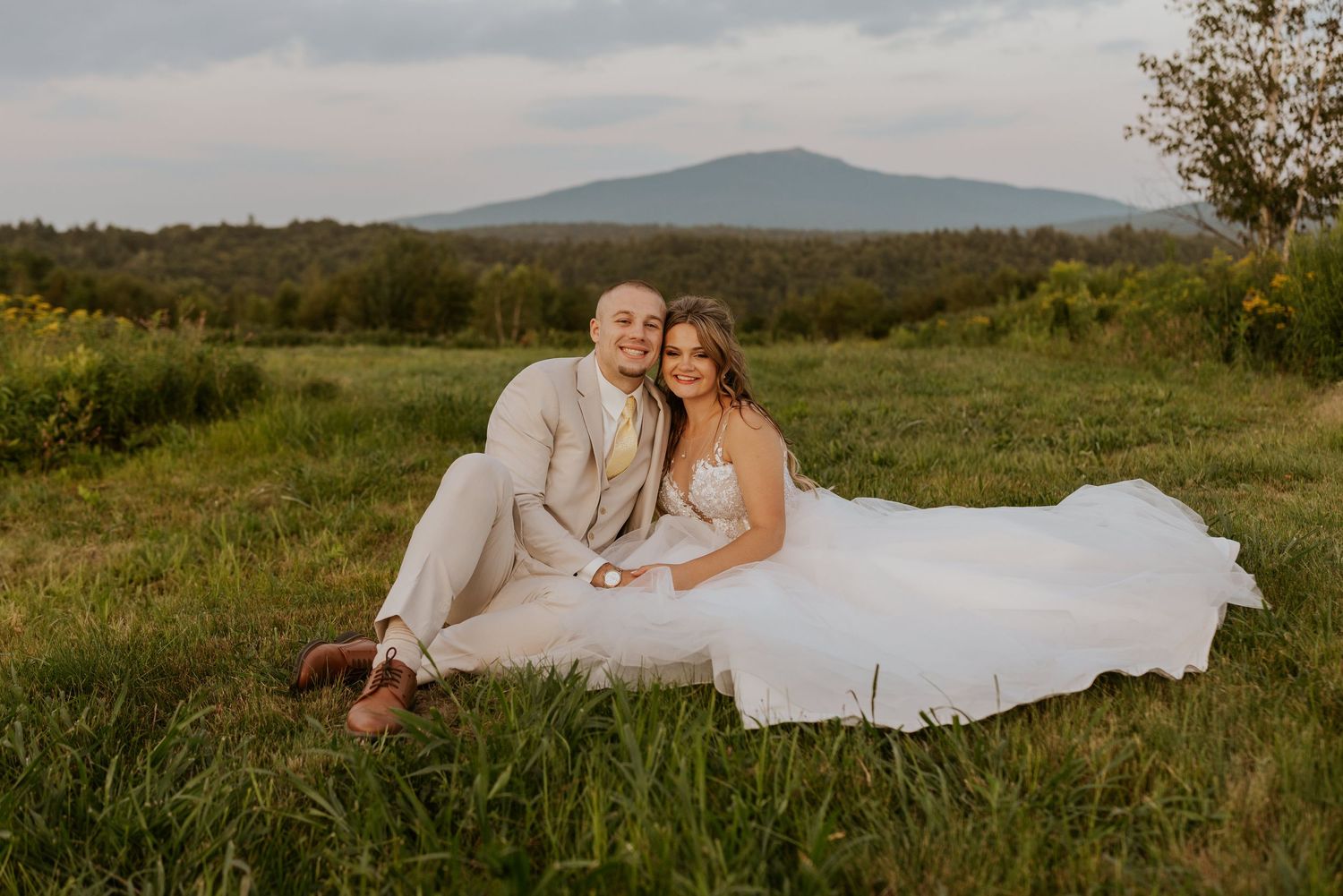 Bride and groom smile together sitting in green meadow with mountain and cloudy sky in the background.