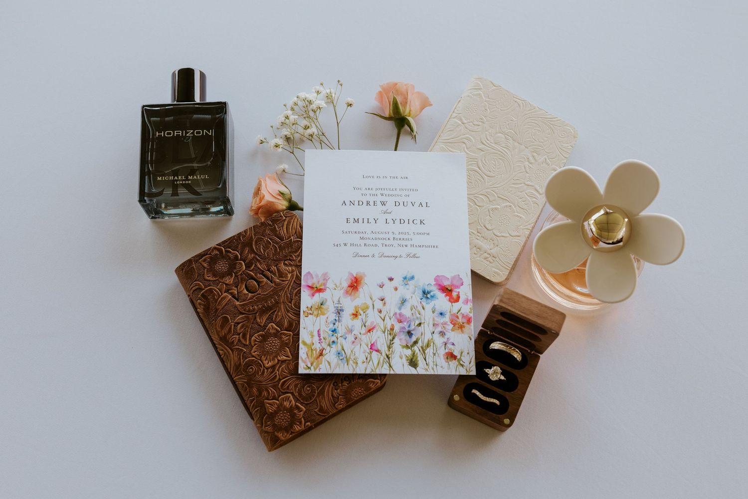 Wildflower wedding invitation flatlay with perfume bottle, leather journal, rings, and floral accents.