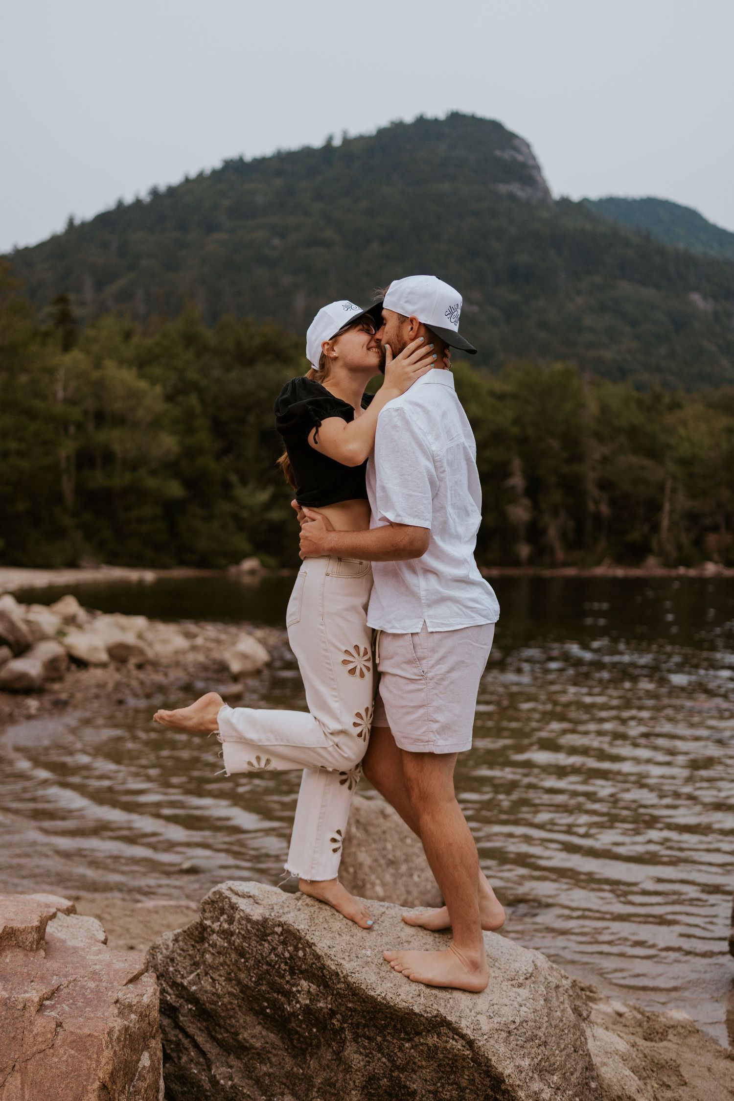 Couple sharing a kiss while standing on rocks by a scenic mountain lake.