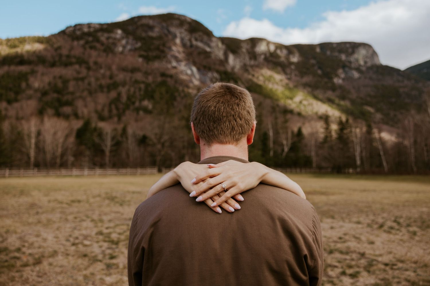 Close-up of a woman resting her hand on a man's shoulder as they face a dramatic rocky mountain landscape.