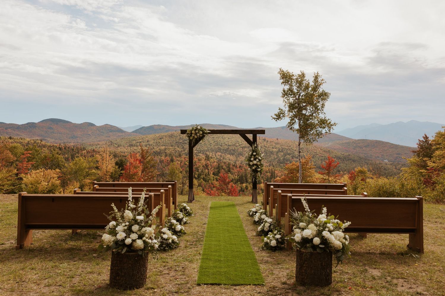 Outdoor fall wedding ceremony setup with wooden pews, green aisle runner, white floral arrangements, and mountain backdrop.