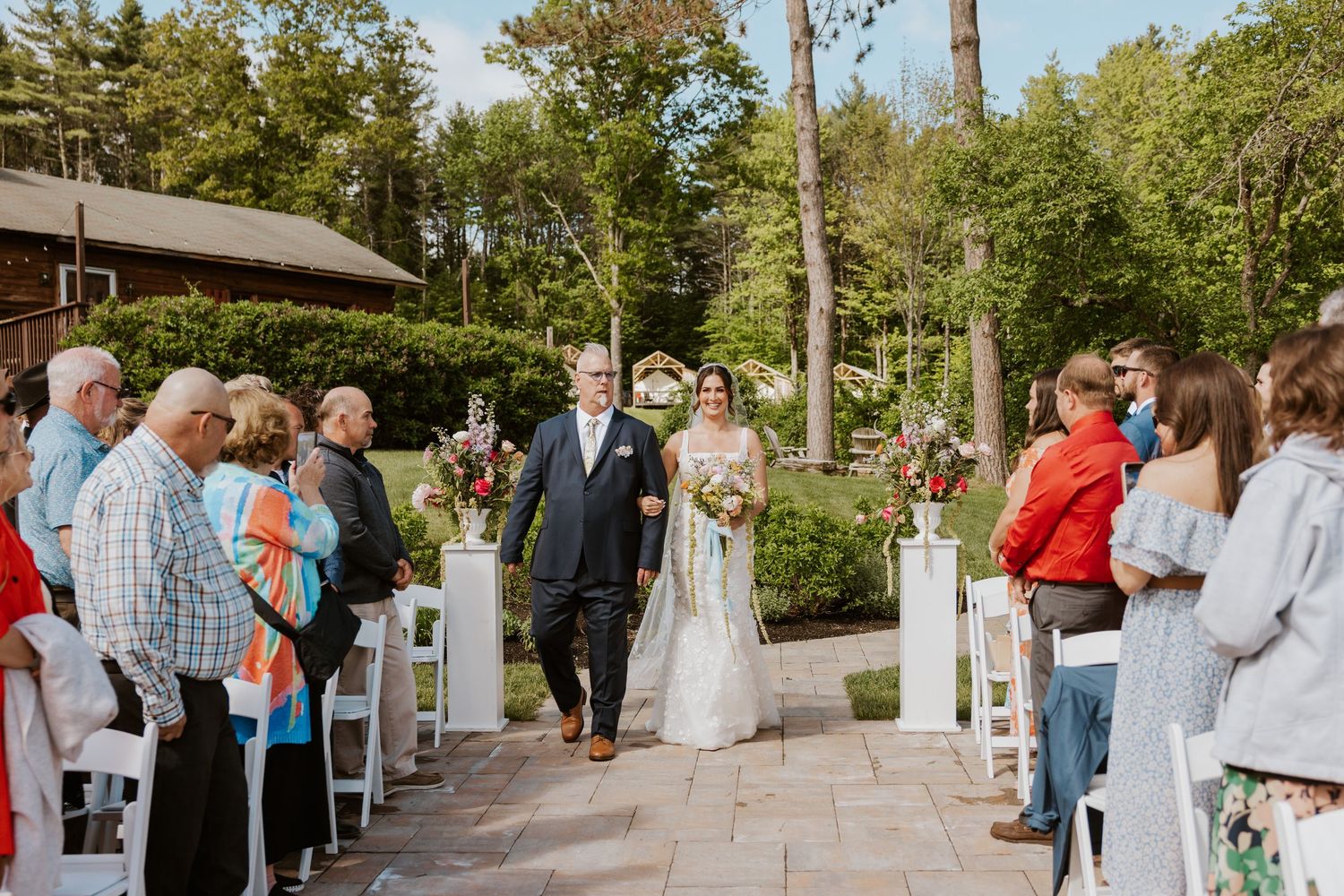 Father escorts bride down outdoor wedding aisle lined with white chairs and floral arrangements.