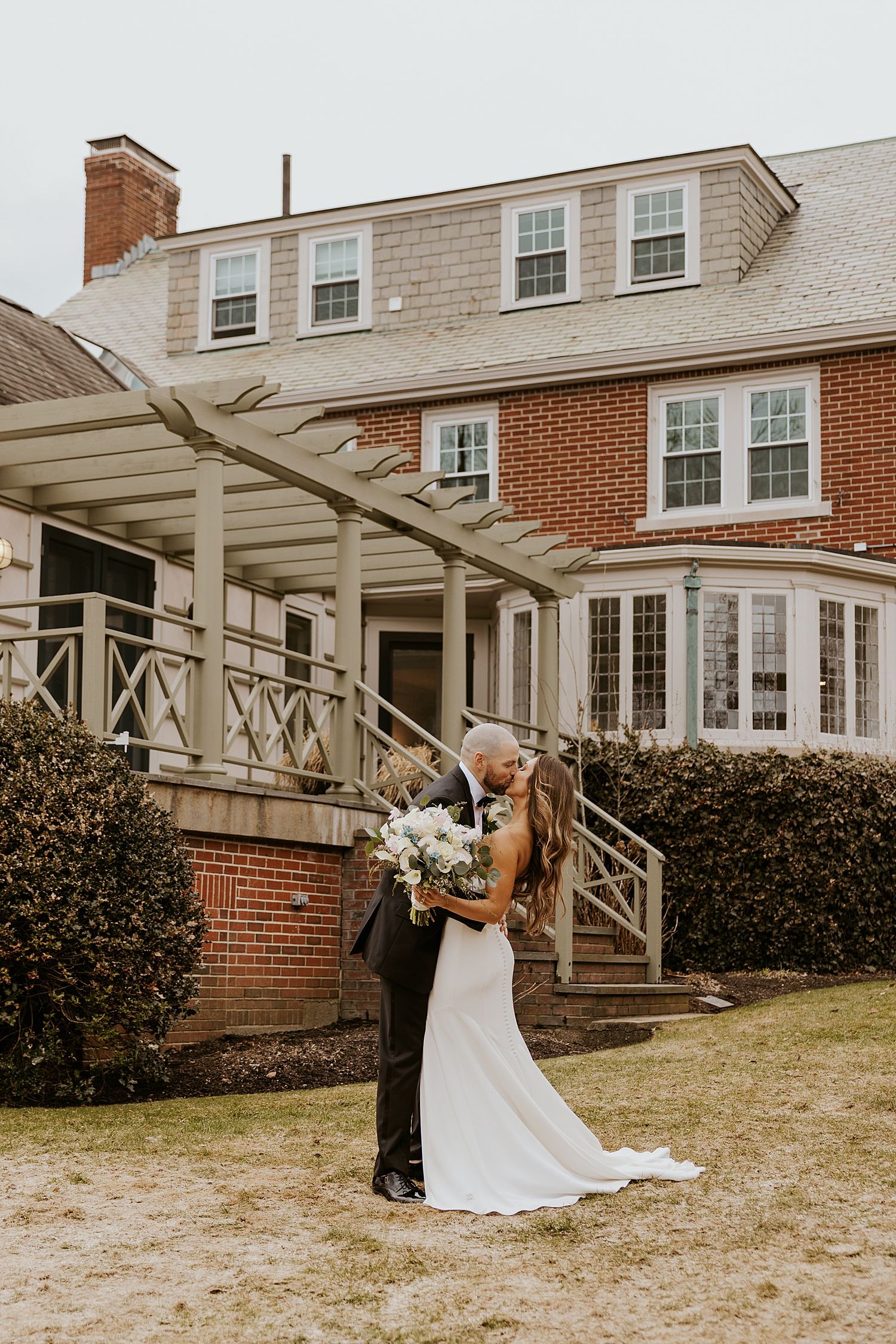 Bride and groom share romantic kiss outside elegant brick estate home with pergola and lush hedges.