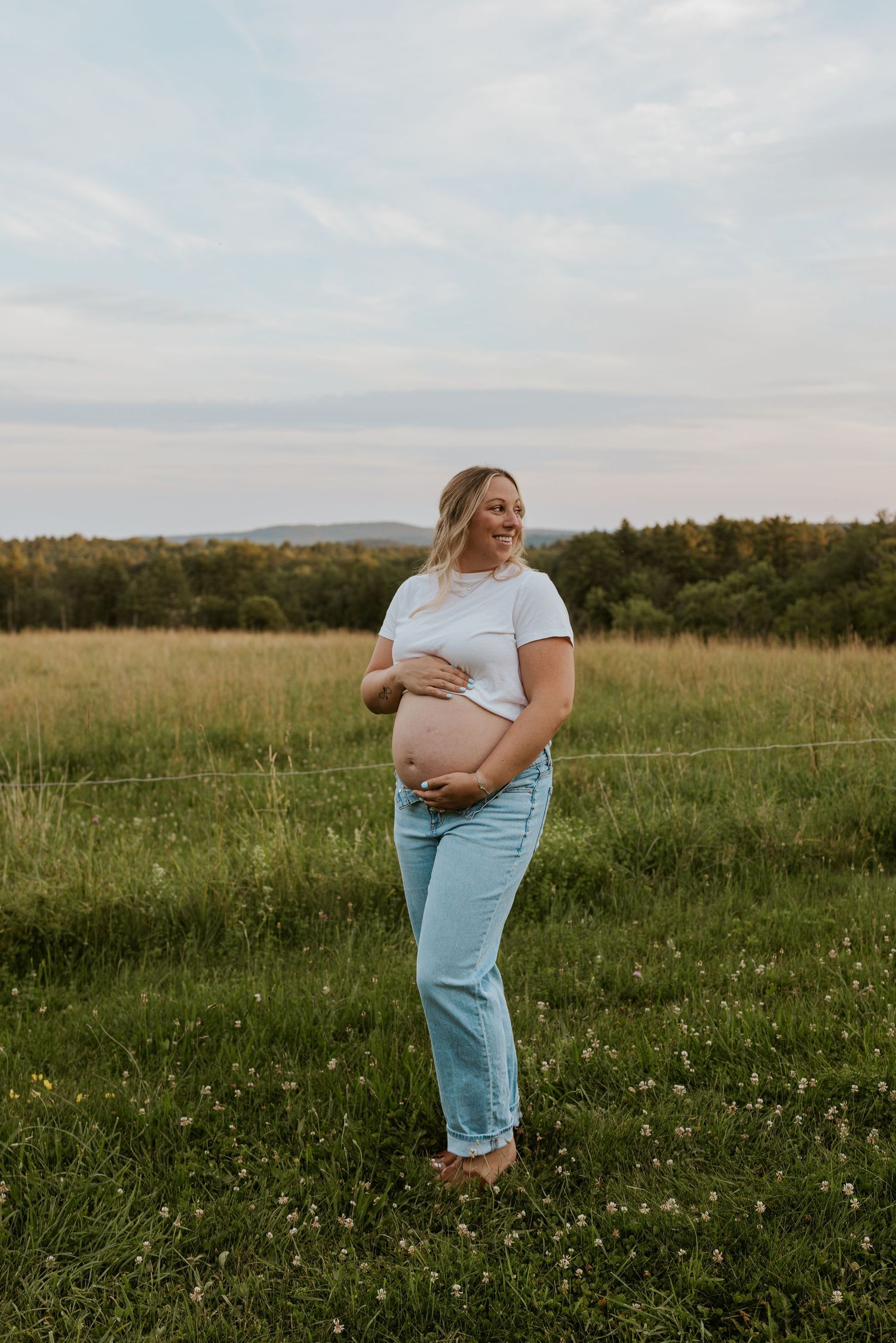 Smiling pregnant woman in a white tee and jeans cradling her baby bump while standing in a sunny green field.