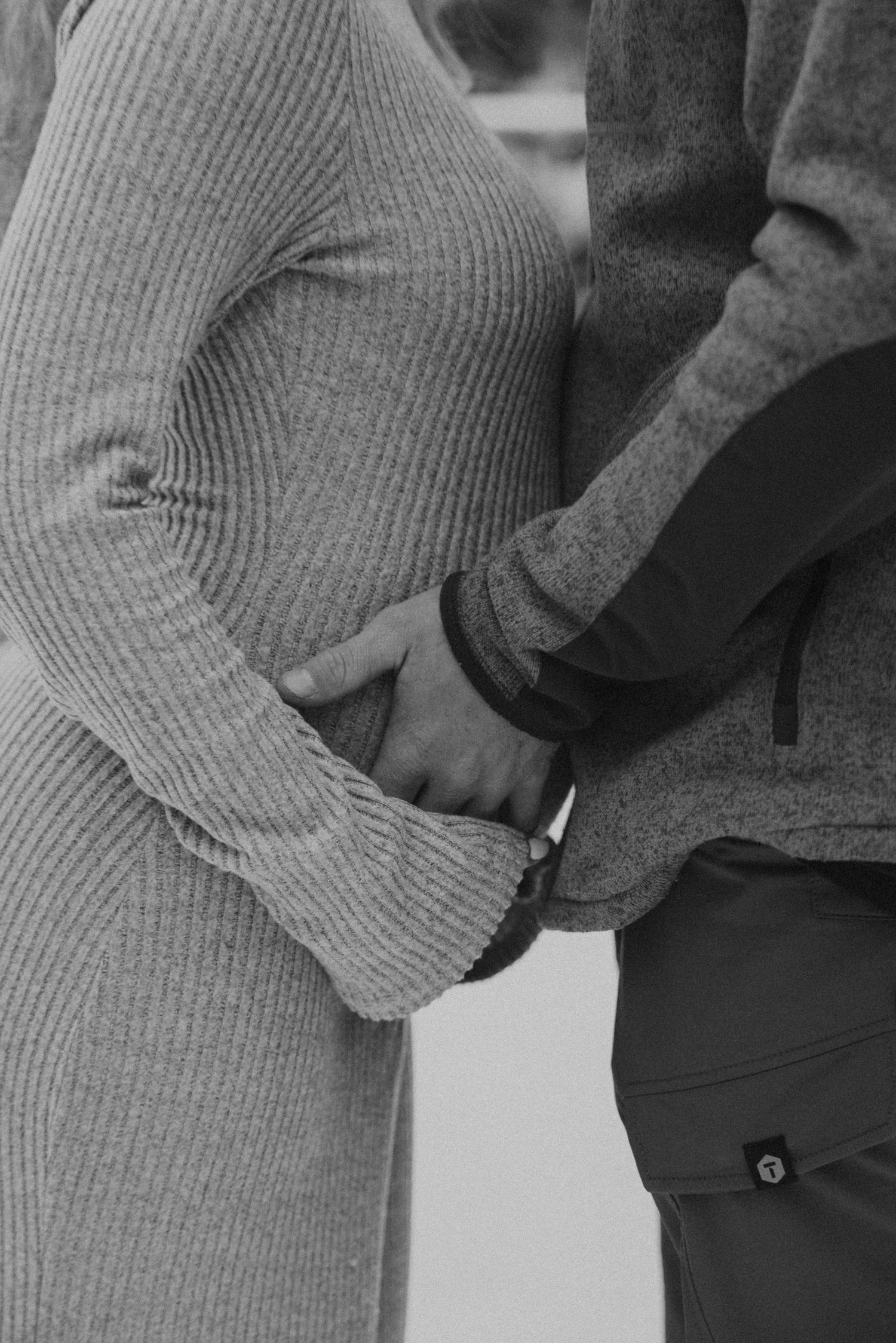 Black and white close-up of couple holding hands over pregnant belly in knit winter dress.