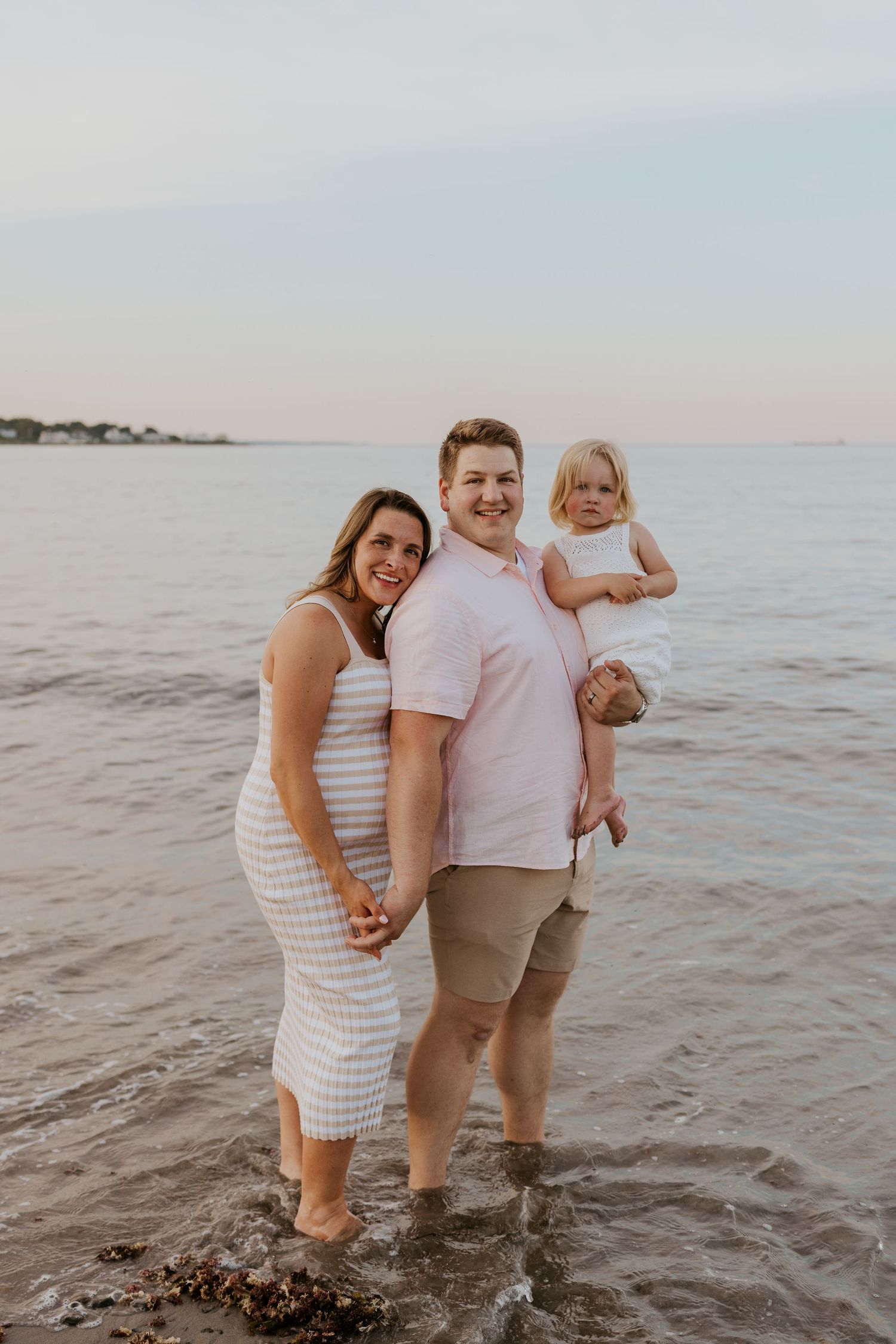 Pregnant woman in striped dress and partner holding toddler stand smiling in ocean waves at sunset.