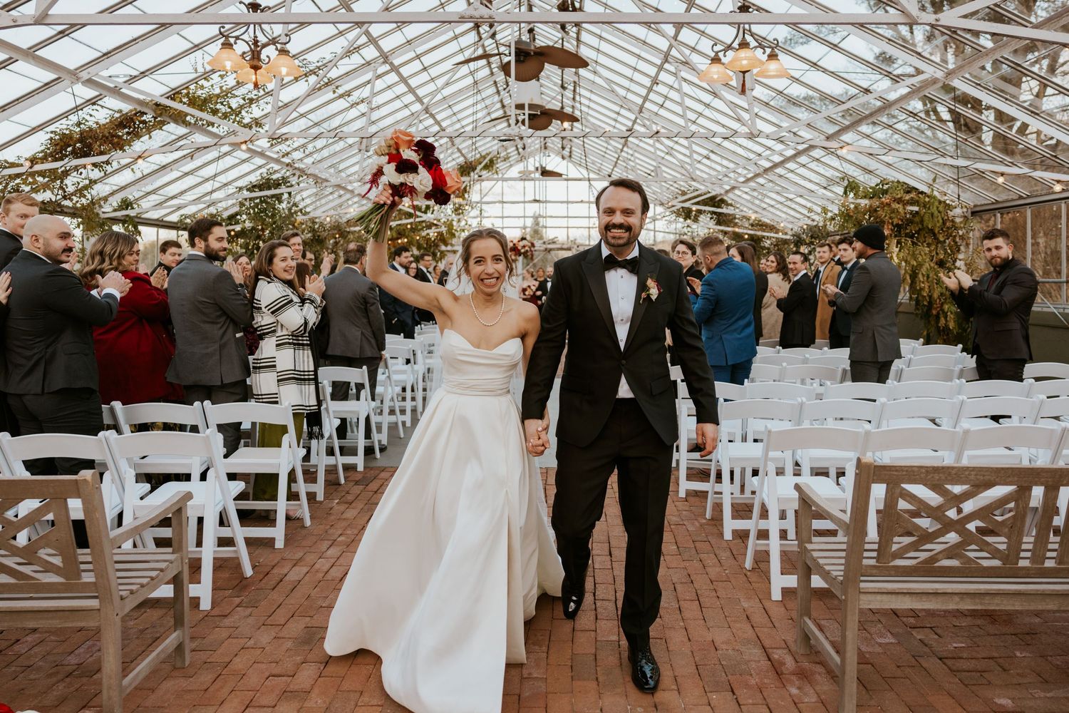 Bride and groom walk joyfully down the aisle in a greenhouse wedding venue with white chairs.