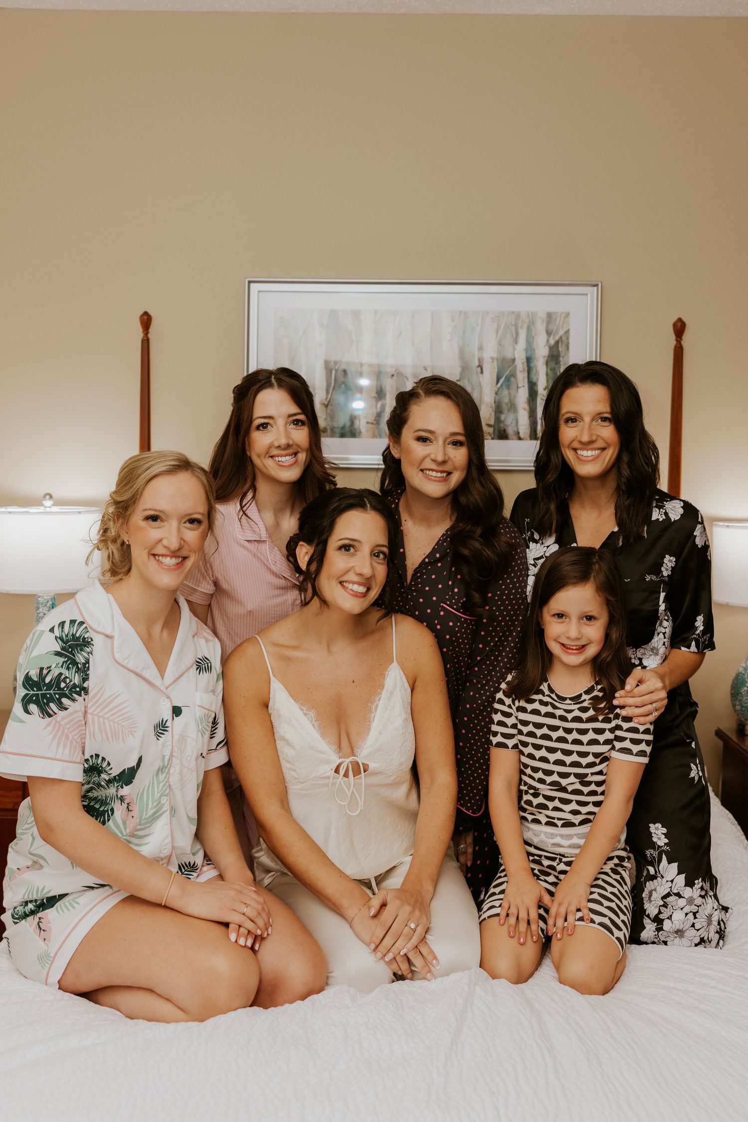 Bride and bridesmaids smiling together on a hotel bed in robes and pajamas during wedding morning preparations.