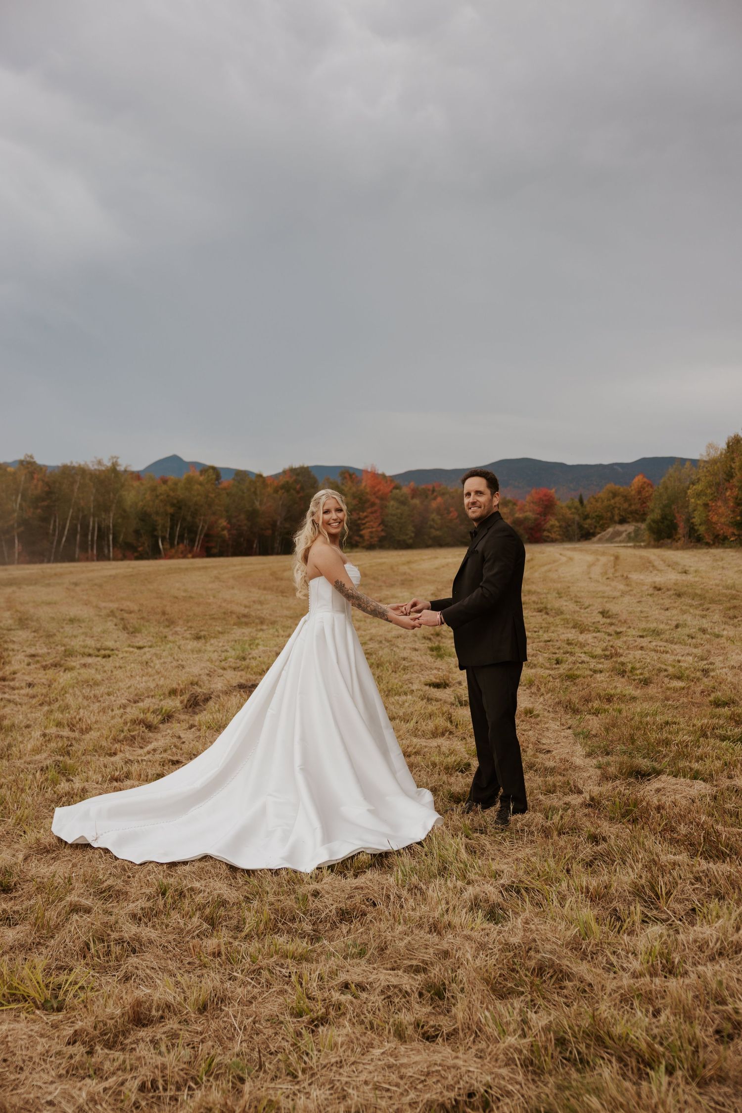 Bride and groom holding hands in golden field with autumn mountains under overcast sky.