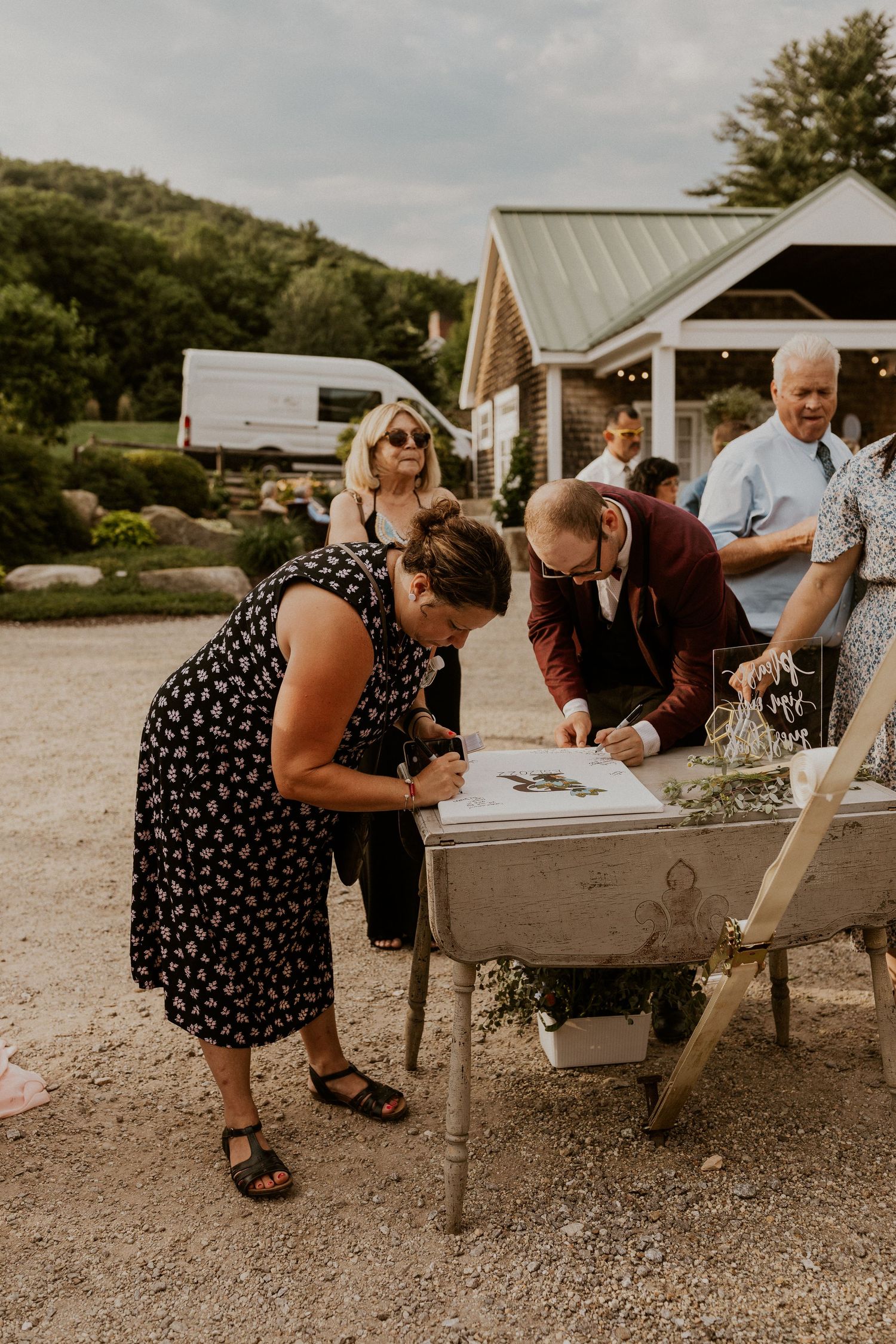 Guests gather around a rustic wheelbarrow guest book at an outdoor wedding venue.