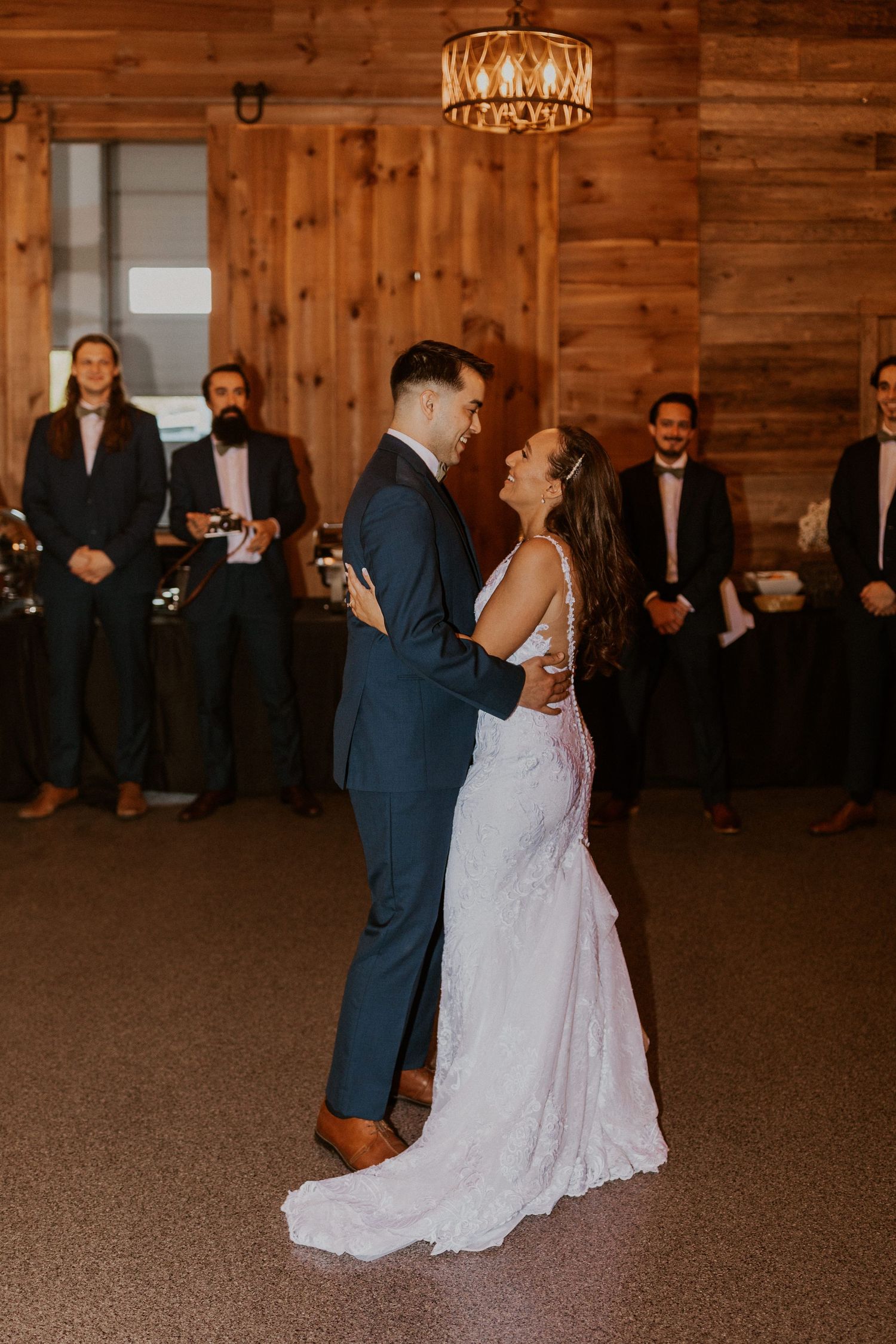 Wedding couple dances beneath elegant chandeliers in a rustic wooden reception hall.