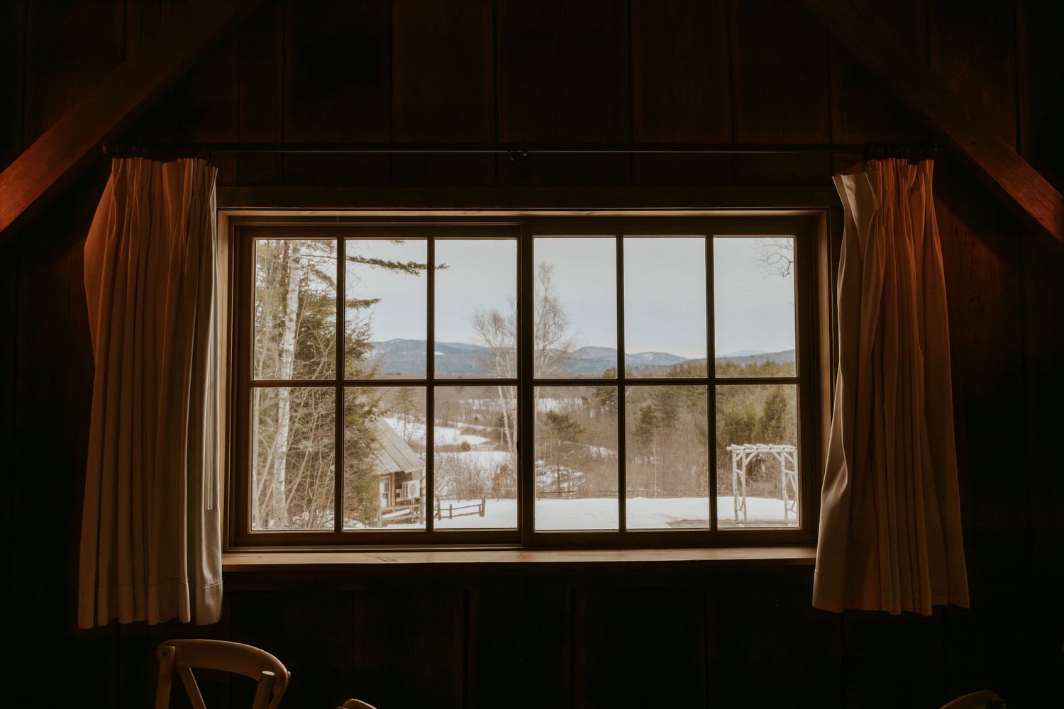 Rustic cabin window framing a serene snowy winter landscape with mountains in the background.