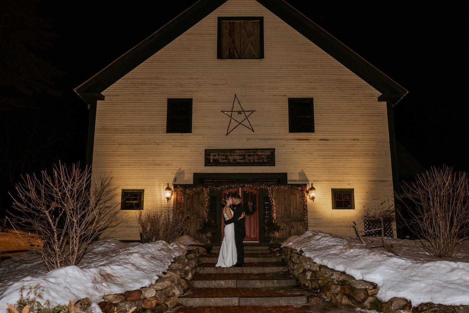 Bride and groom kissing on steps of illuminated white barn wedding venue at night in winter snow.